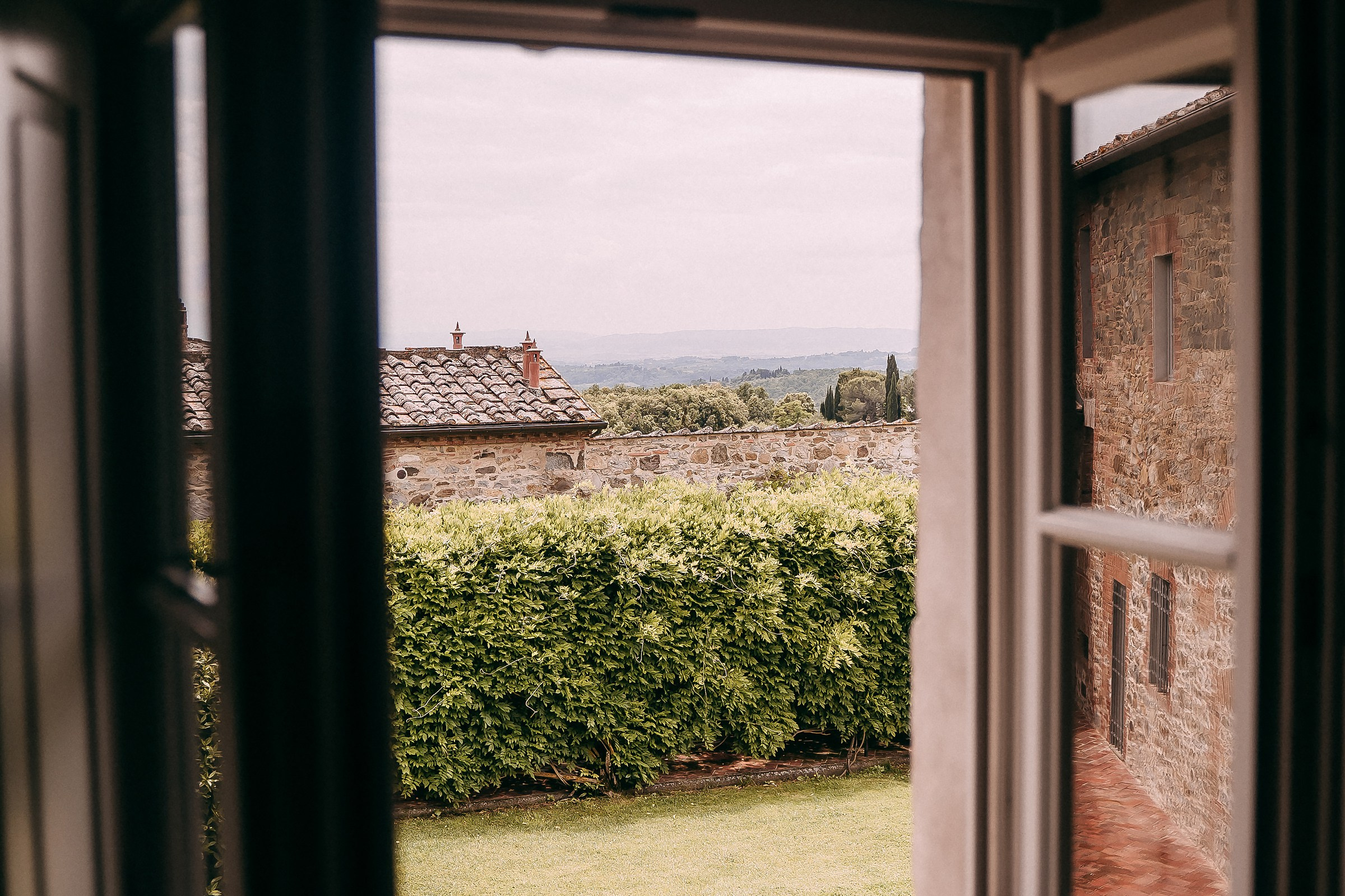 Charming Tuscan countryside seen through a window, revealing rustic stone buildings and lush green hedges.