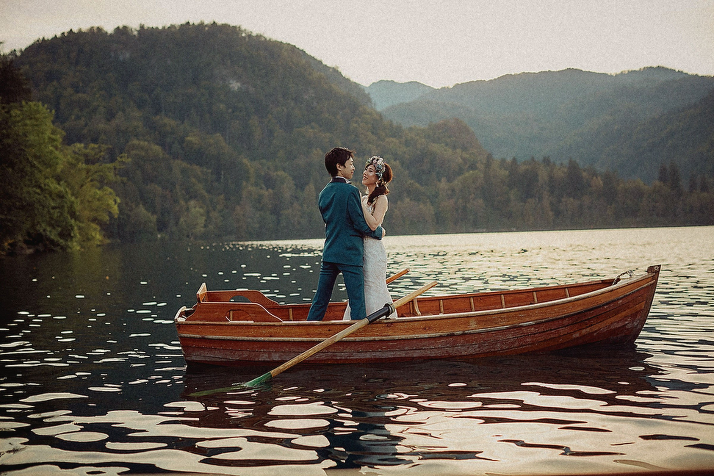 A Japanese bride wearing a floral headpiece and her groom stand upright in a rowboat as they enjoy their time on a lake.