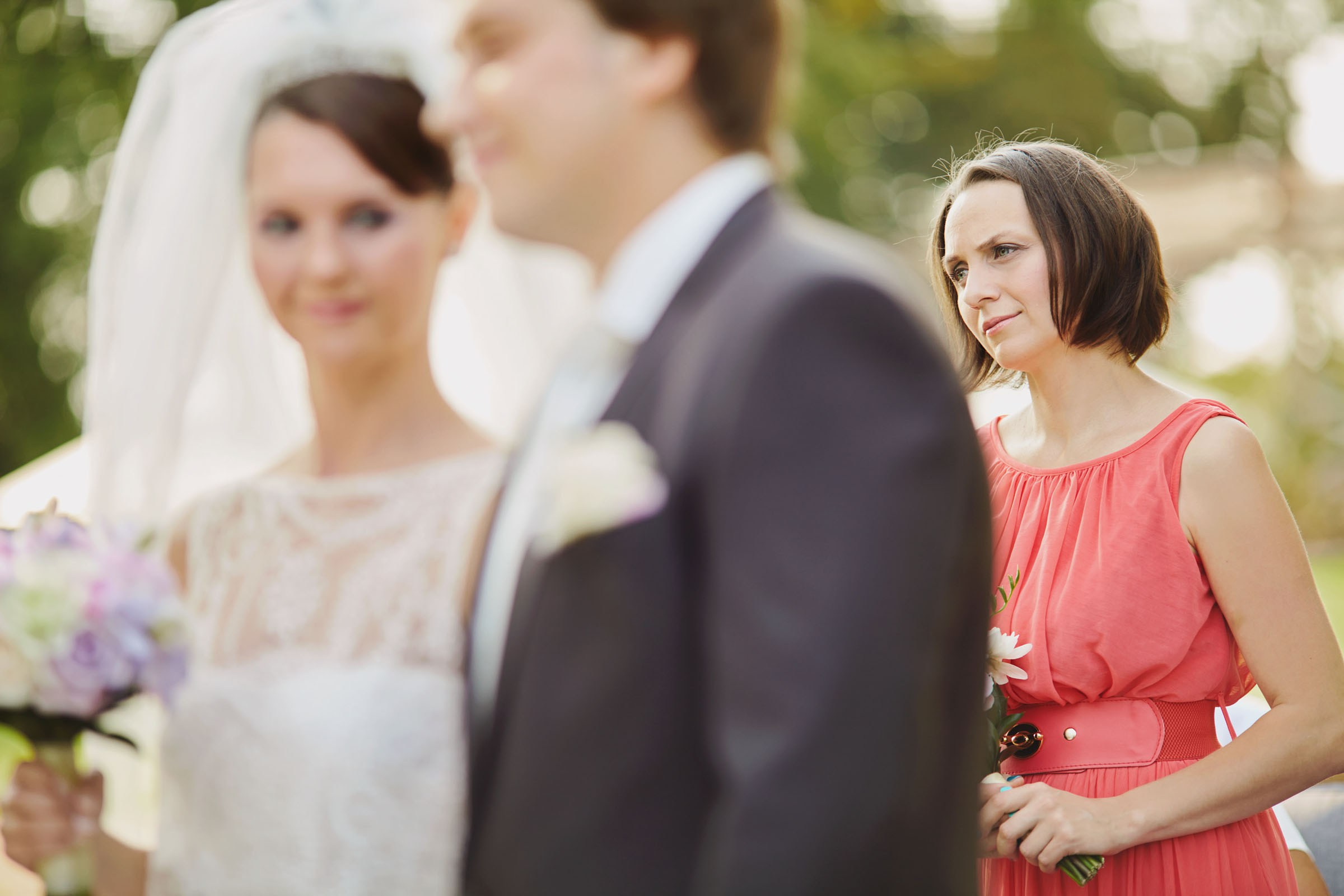 The aunt of the bride holding a bouquet of flowers stands by as the bride and groom listen to the wedding officials during their outdoor wedding.