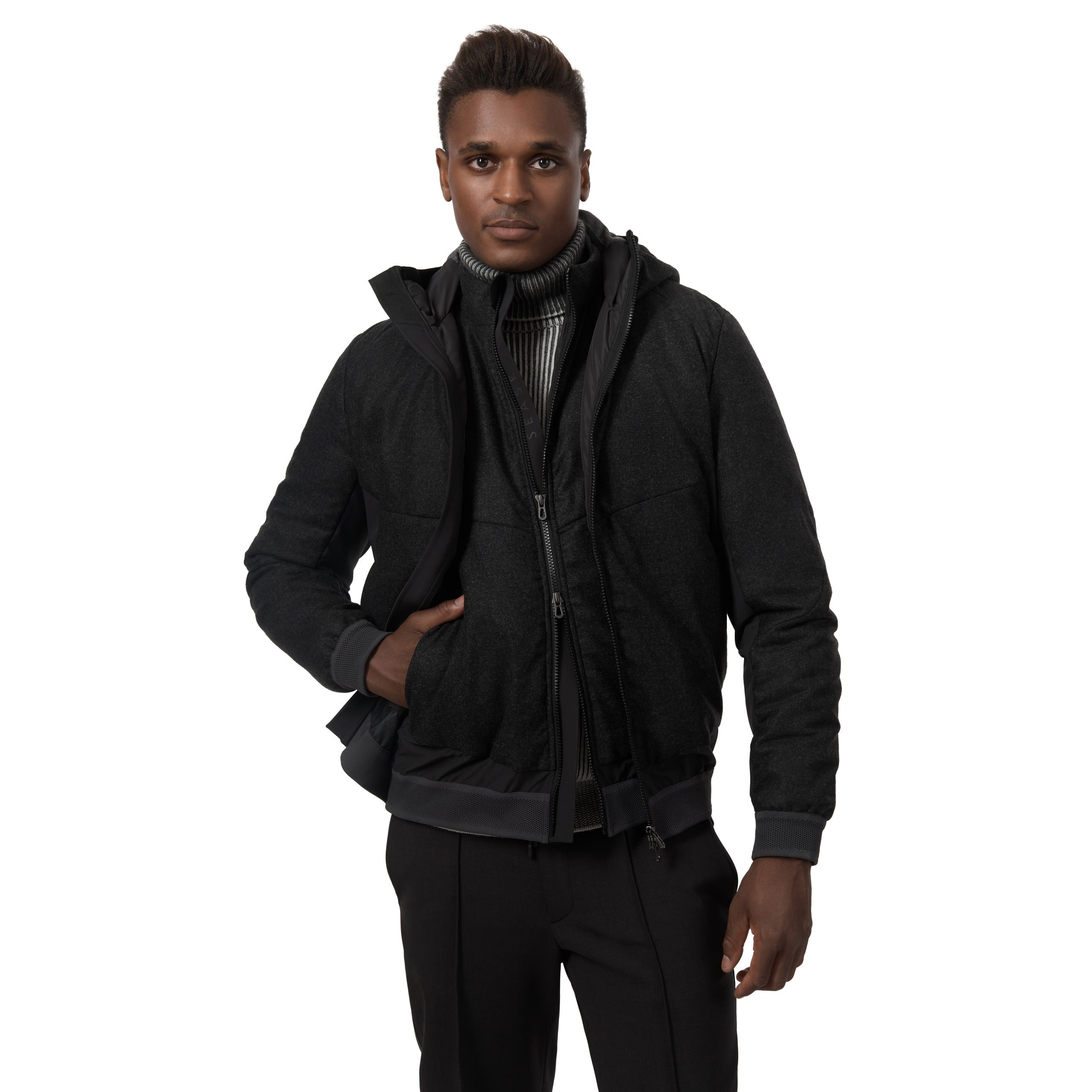 Model in schwarzer Weste und grauem Pullover, Studioaufnahme auf weißem Hintergrund.