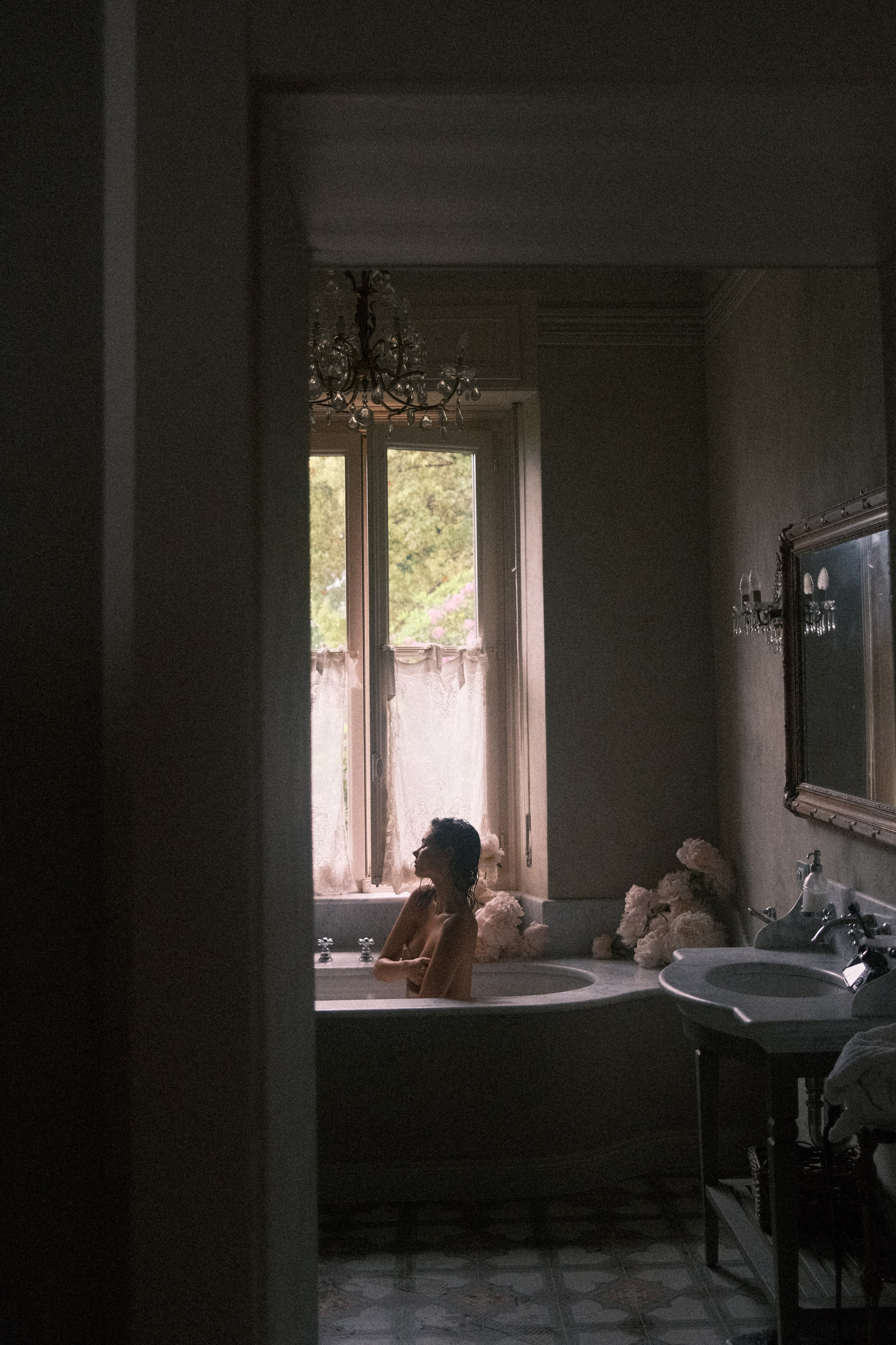 A moody shot capturing a dimly lit bathroom, with the silhouette of a woman relaxing in a clawfoot bathtub by a large window draped with lace curtains.
