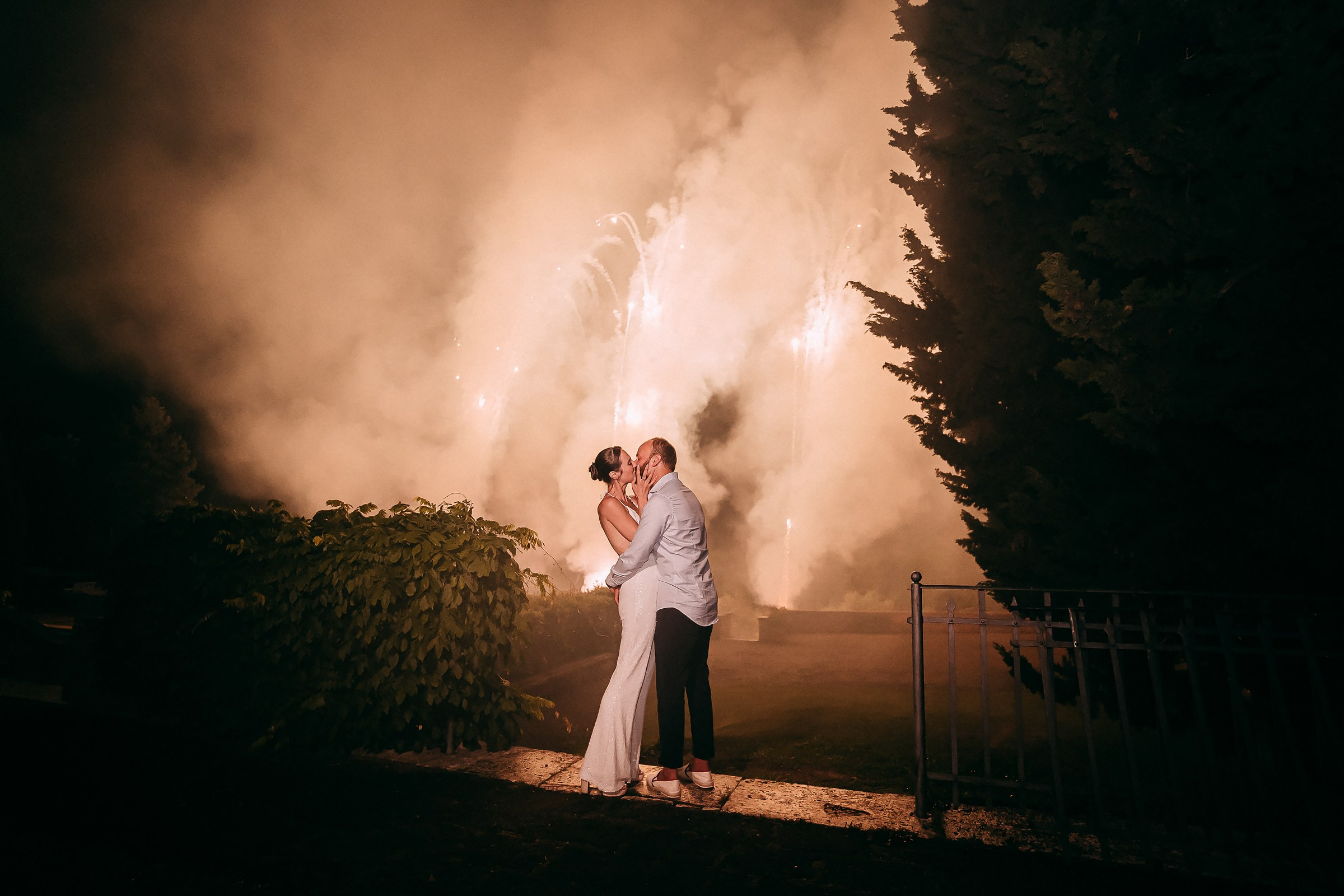 A newlywed couple embraces passionately, surrounded by the enchanting glow and smoke of a magnificent nighttime fireworks display.