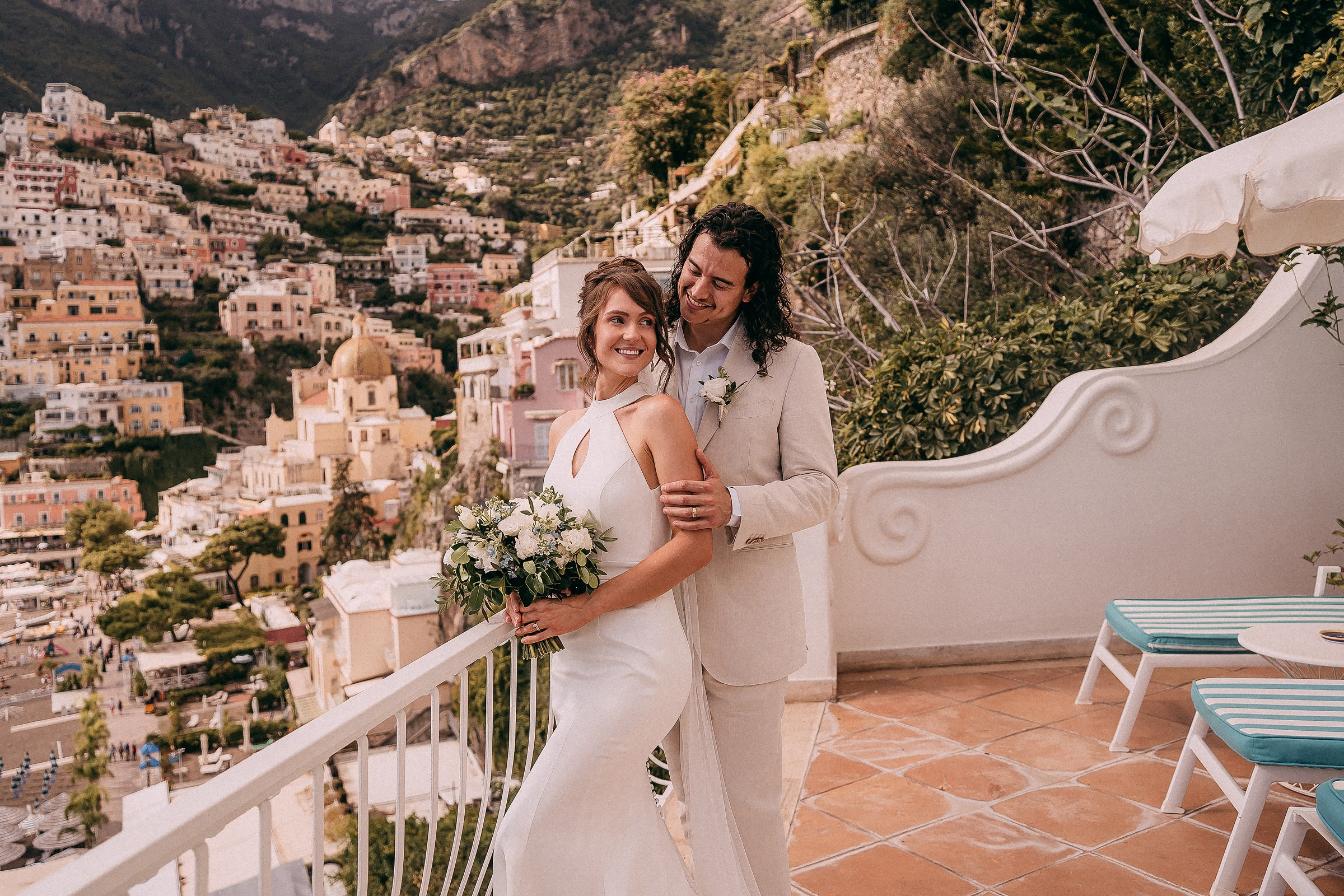 A joyful bride and groom standing together on a terrace in Positano, the bride holding a bouquet of white flowers, surrounded by a stunning view of the Amalfi Coast.