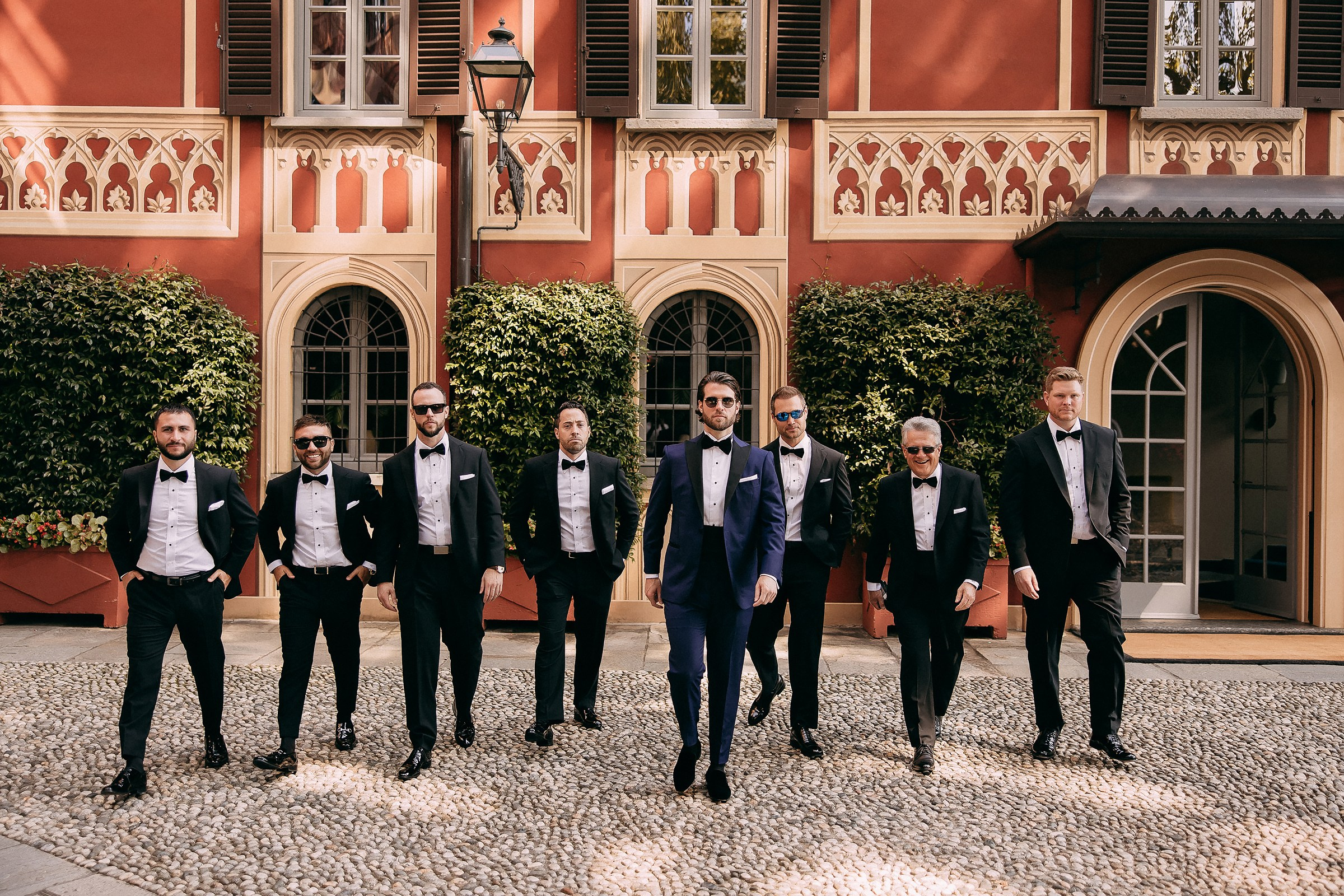 The groom and his groomsmen, elegantly attired in tuxedos, stand together in front of a magnificent mansion.