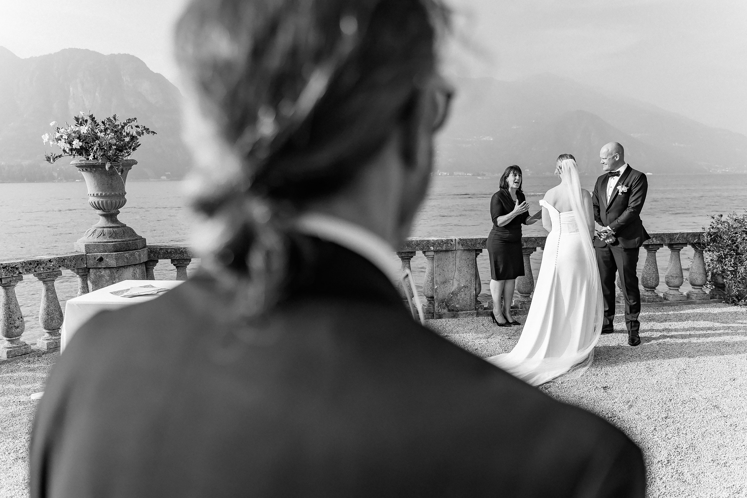 A black-and-white perspective from a guest’s view, showing the bride, groom, and officiant against a stunning lake Como and mountain backdrop.