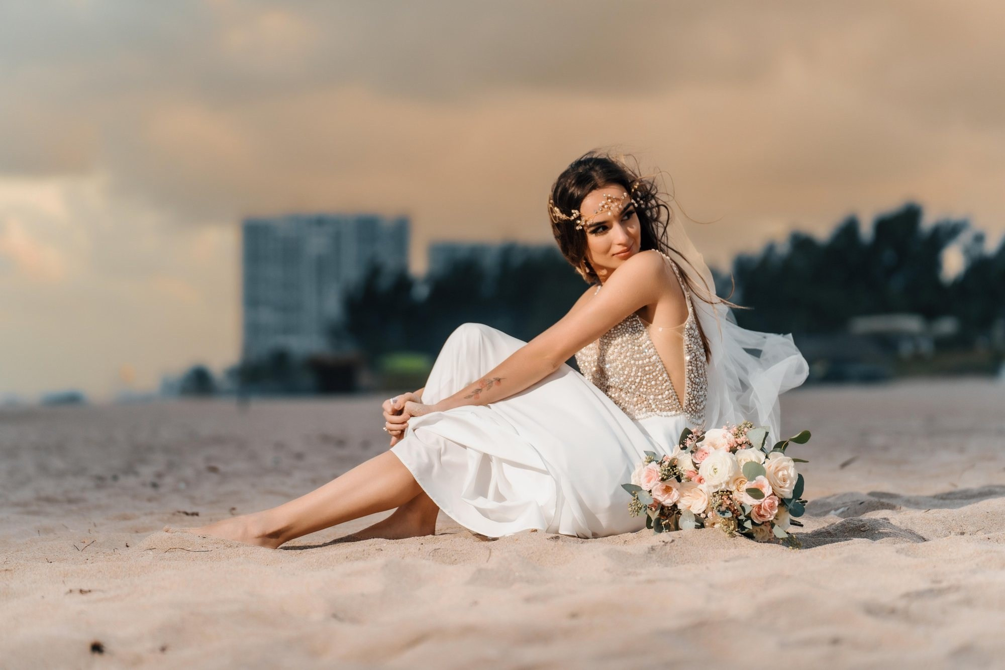 Fort Lauderdale beach wedding photographer — bride sitting on the sand with bouquet, skyline behind