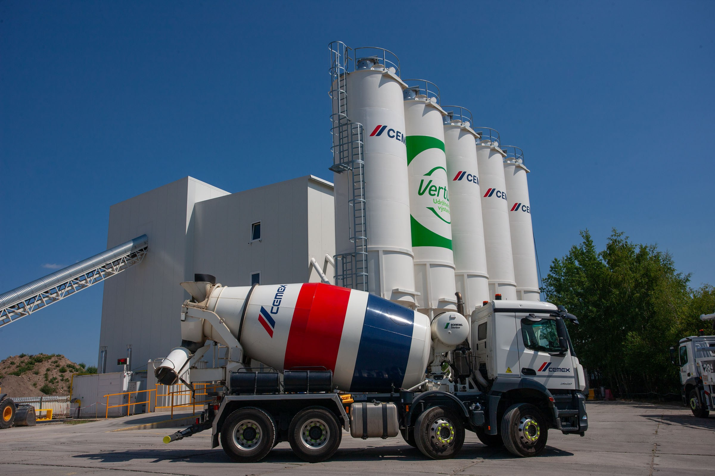 A cement truck is seen against the cement silos as a deep blue sky accentuats the red and whites of the truck.