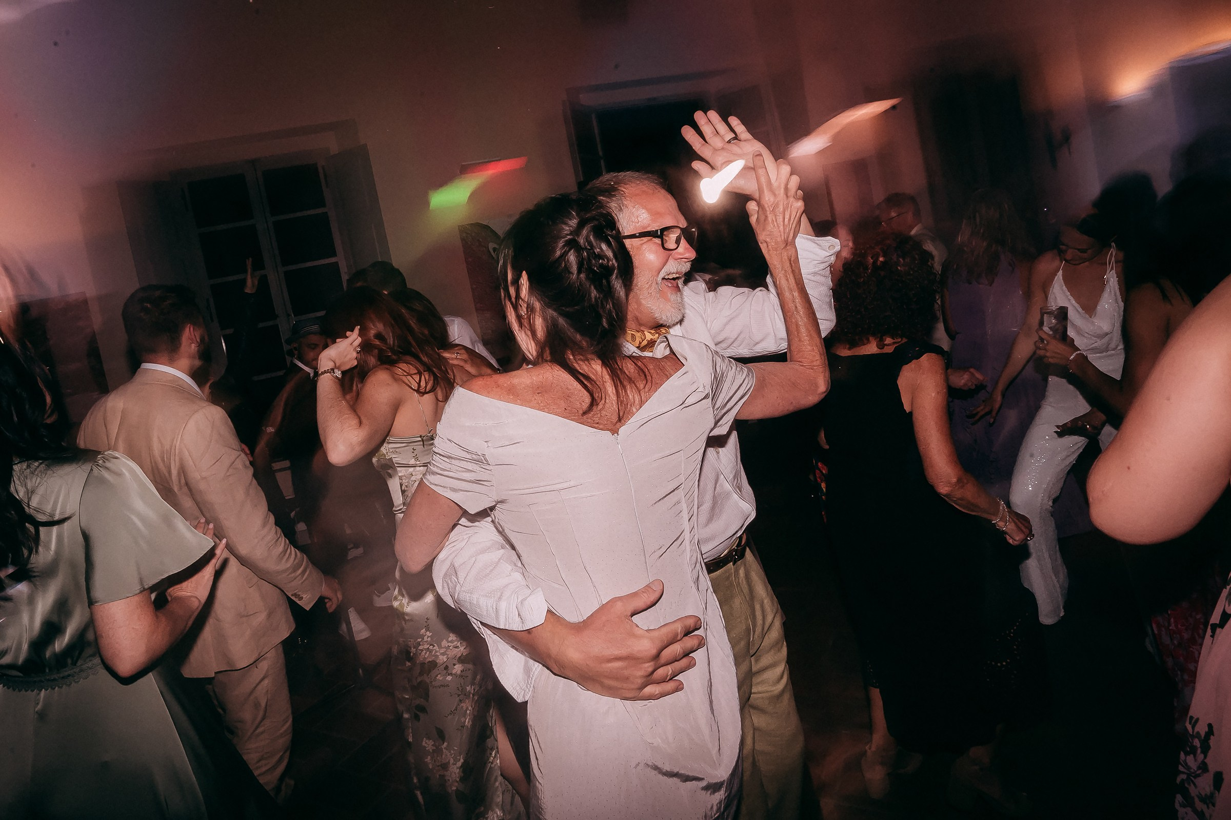 A lively dance floor scene with a couple in focus; the man holds the woman close as she raises her hand in an animated gesture, surrounded by colorful lights and other dancing guests.