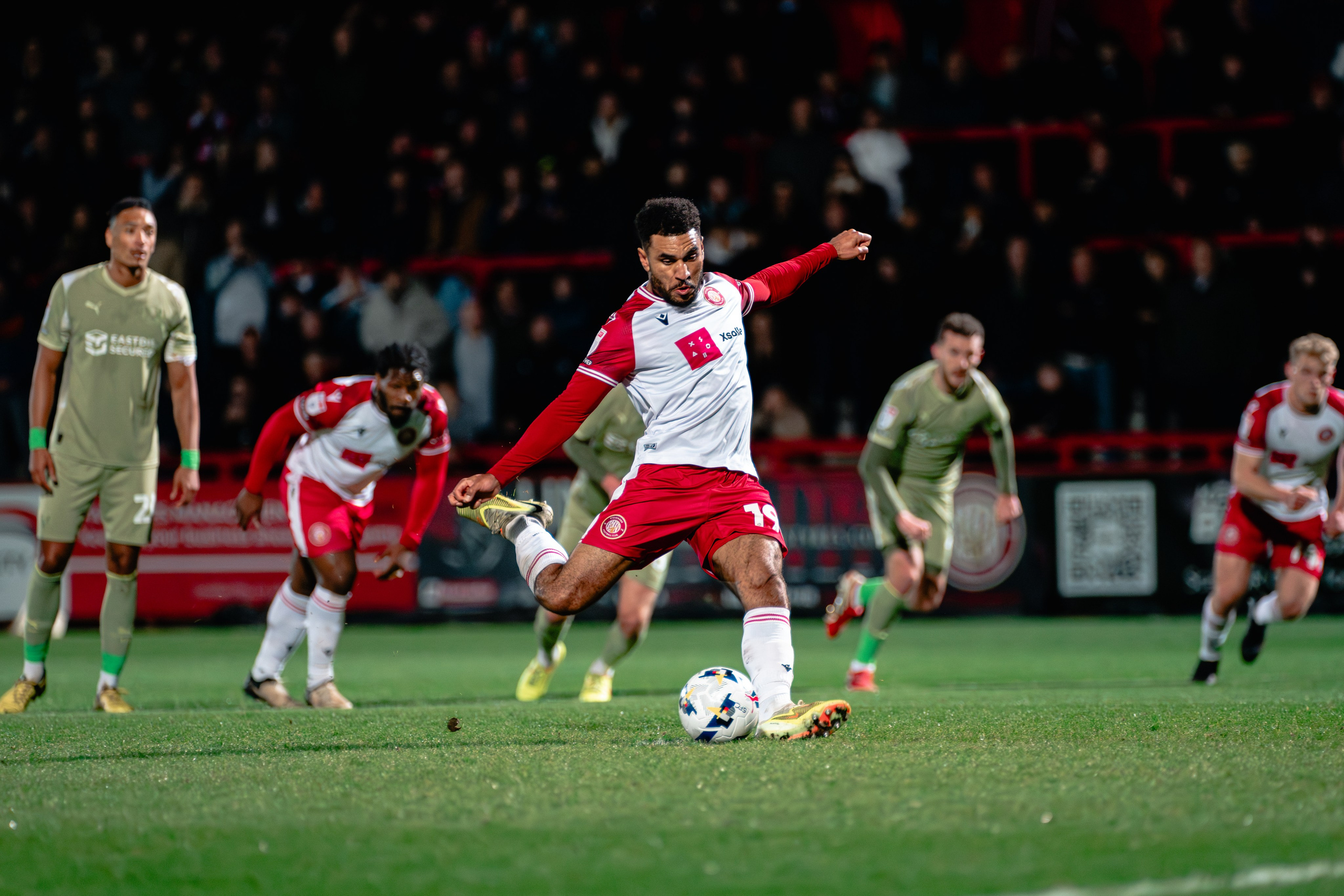 Stevenage player striking the ball during the League One fixture against Leyton Orient under the floodlights.