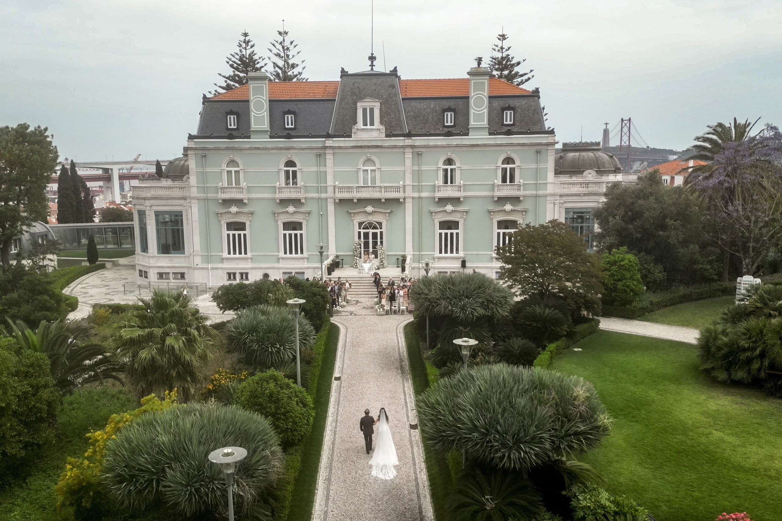 Destination wedding at Pestana Palace Lisbon with the couple walking toward the ceremony and the 25 de Abril Bridge in the background