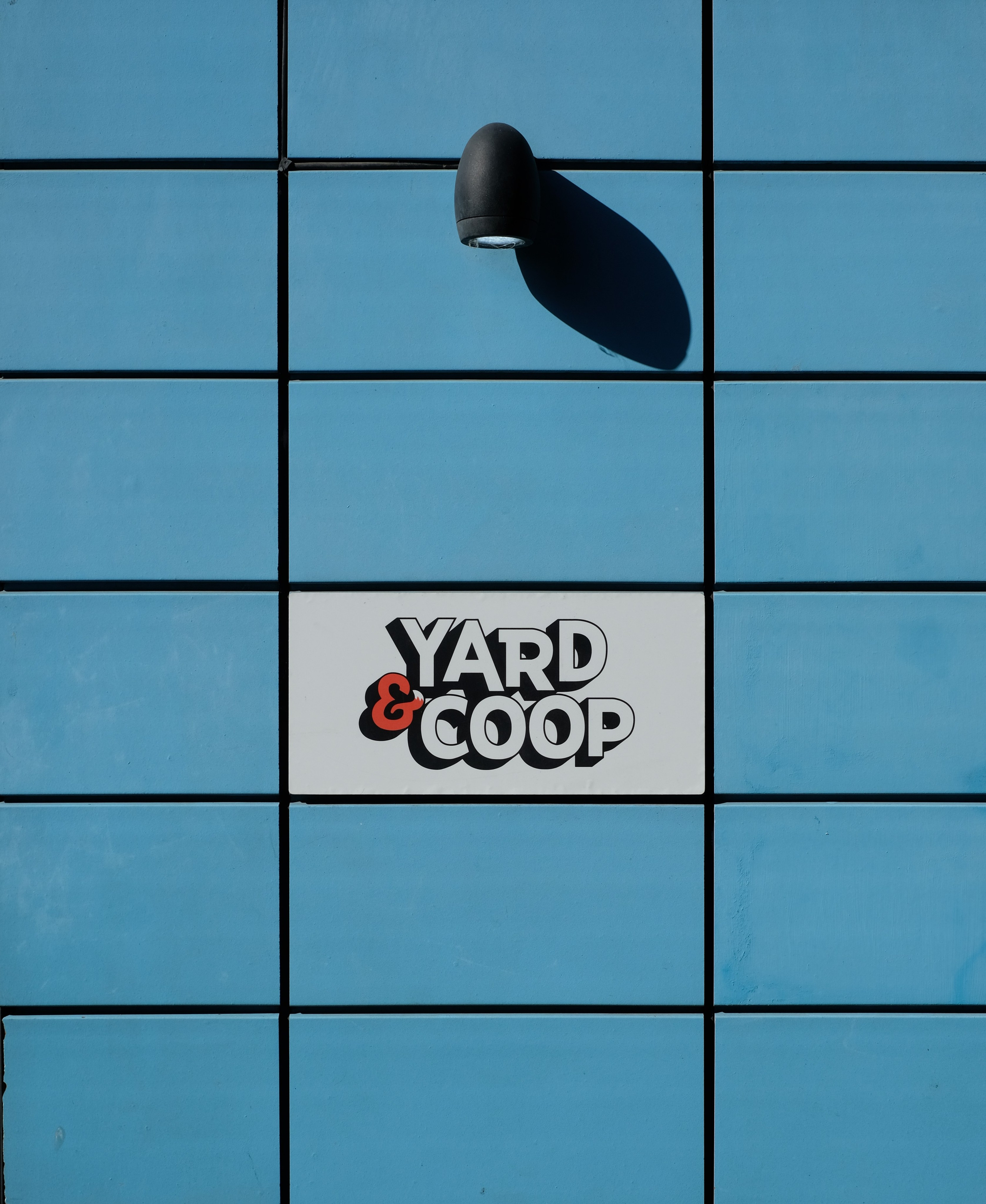 Yard and Coop sign on a blue tiled wall, with a lamp casting a shadow.