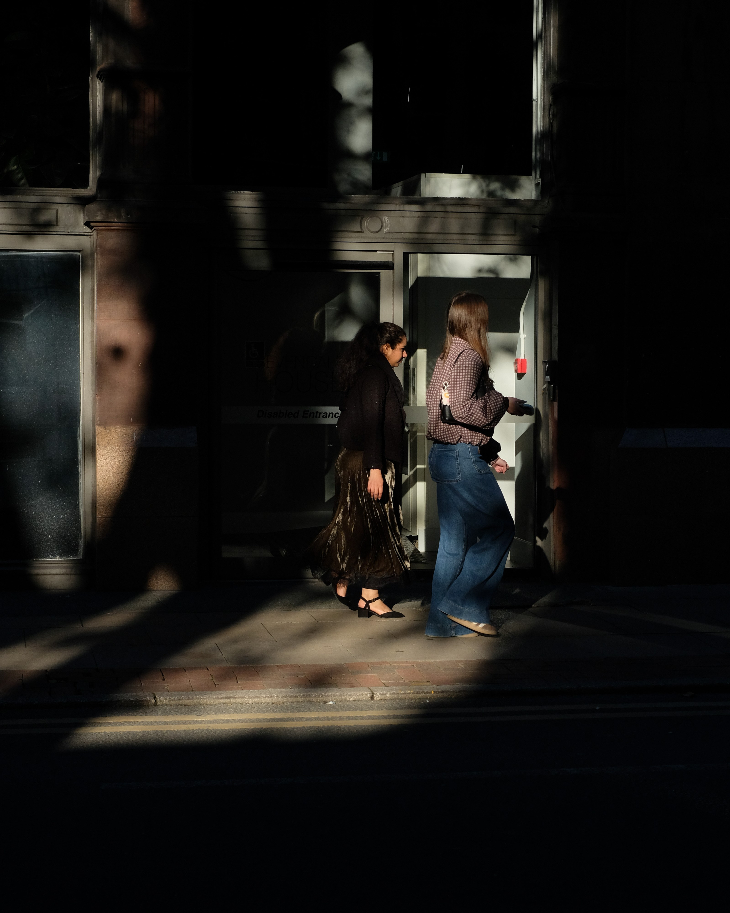 Two pedestrians walking through a shaft of sunlight between deep building shadows.
