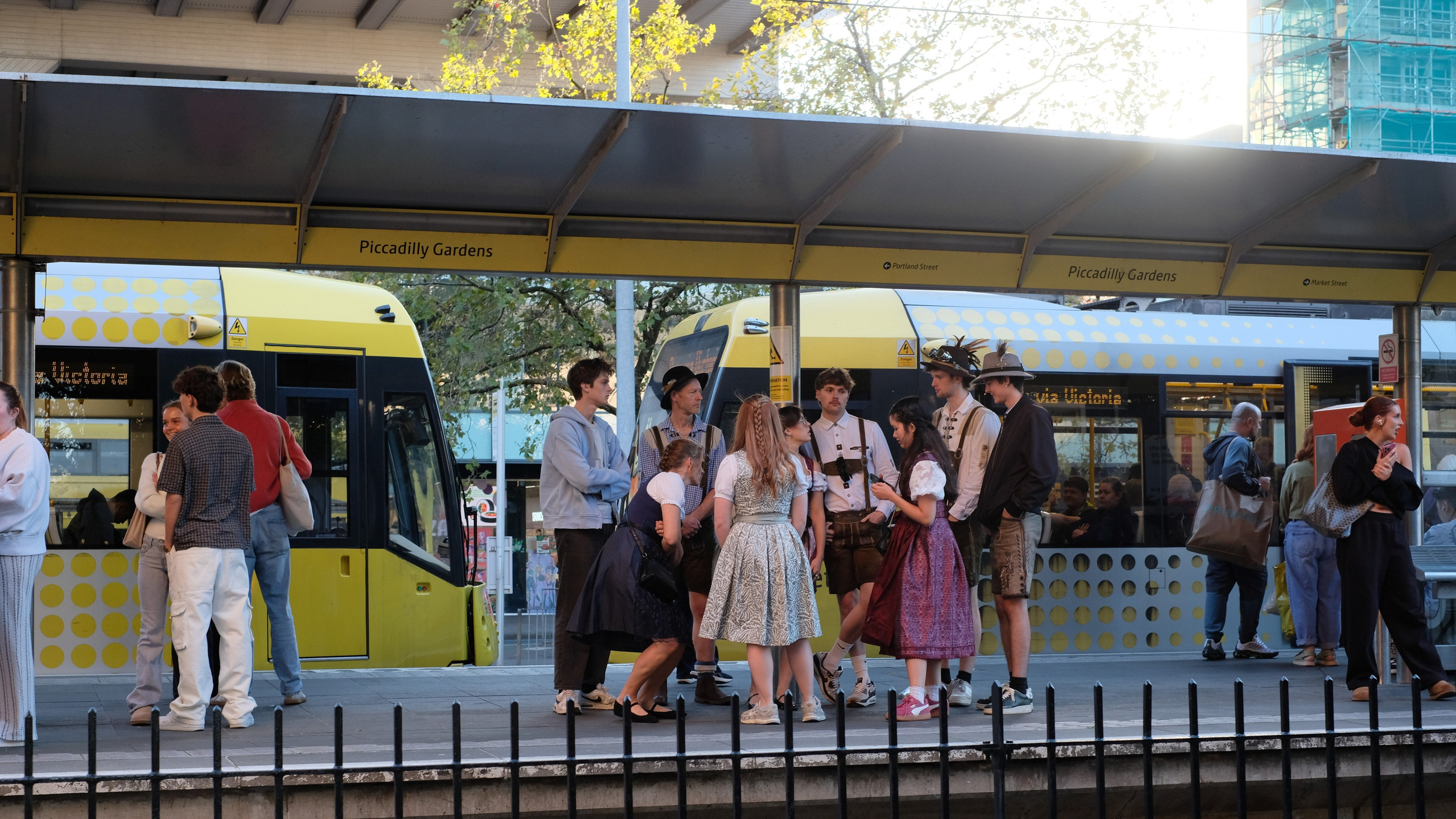 People waiting beside yellow trams at Piccadilly Gardens in Manchester.