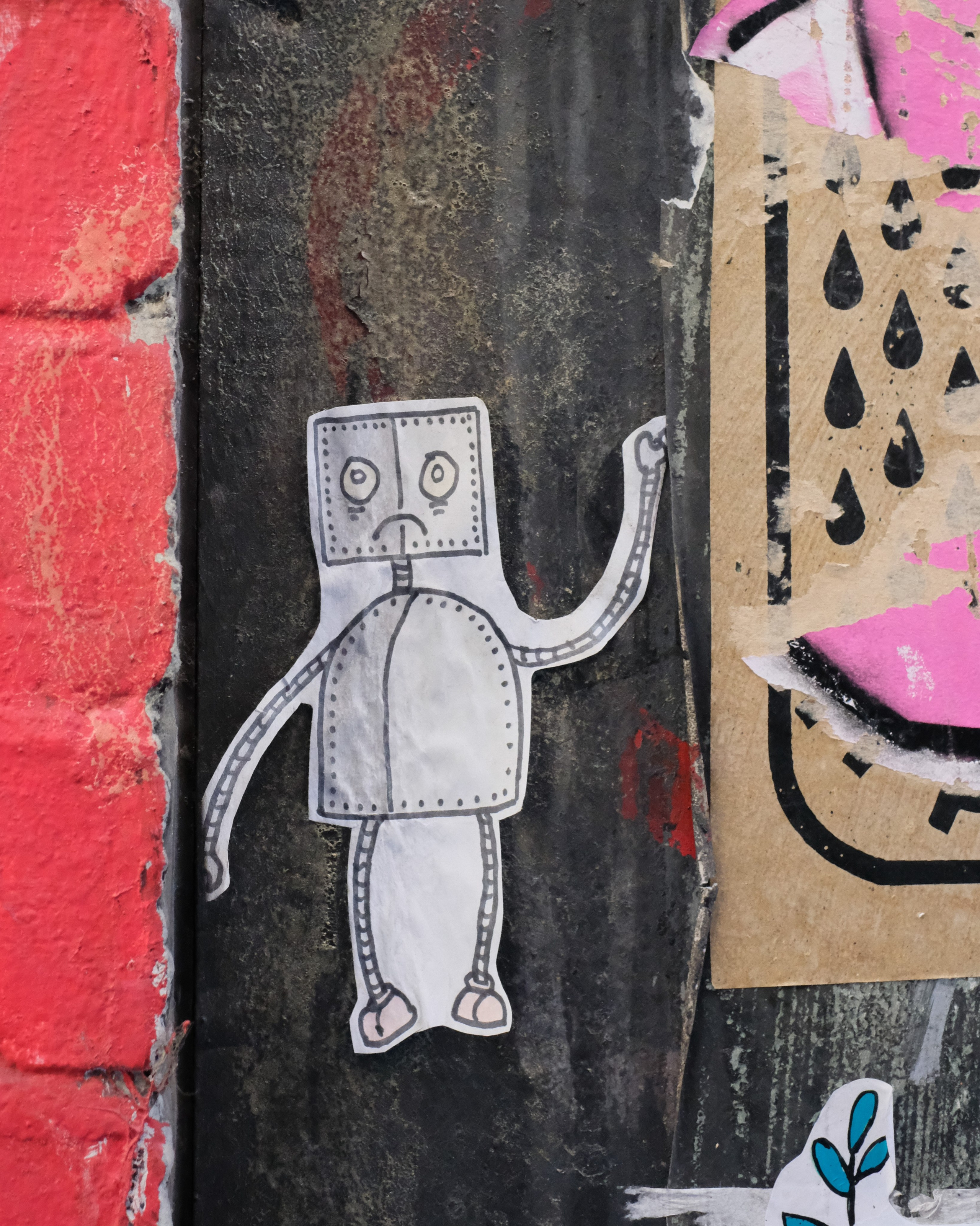 A small paper robot pasted onto a textured wall beside red brick and pink artwork.