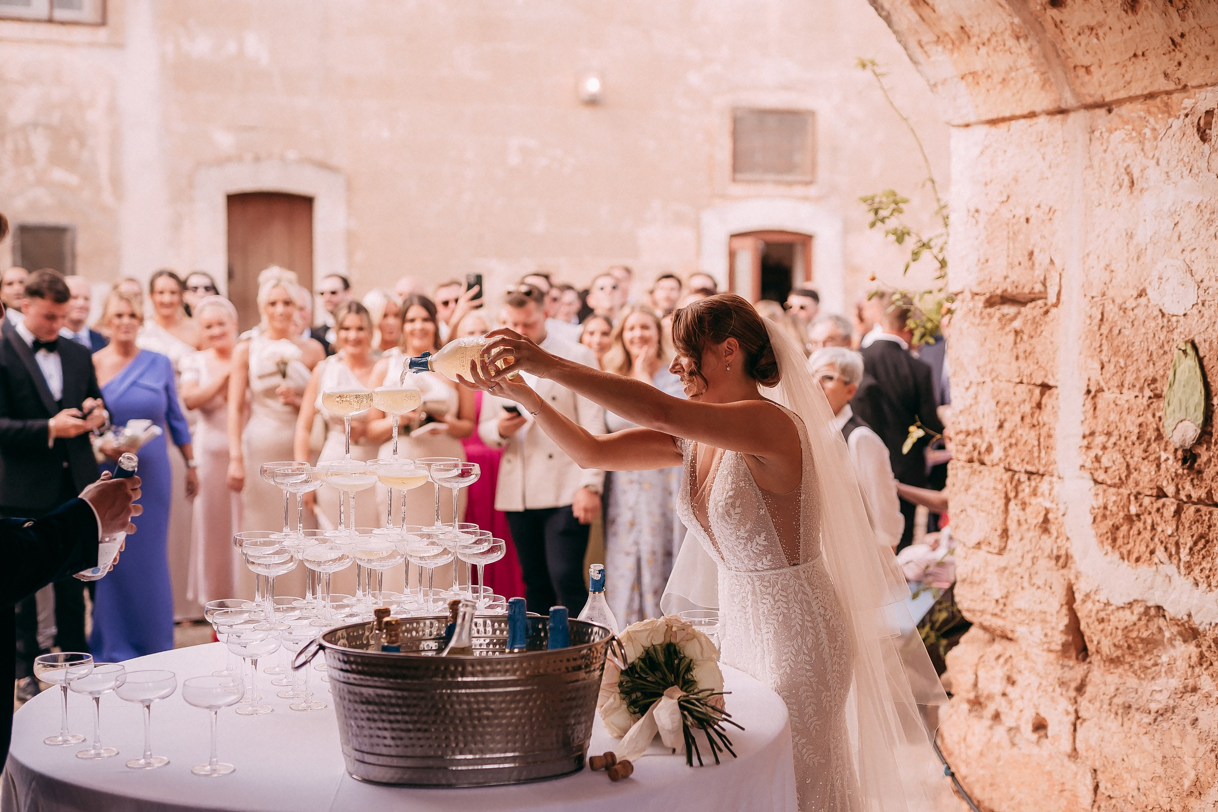 The bride pouring champagne into a cascading tower of glasses, surrounded by excited guests capturing the celebratory moment.