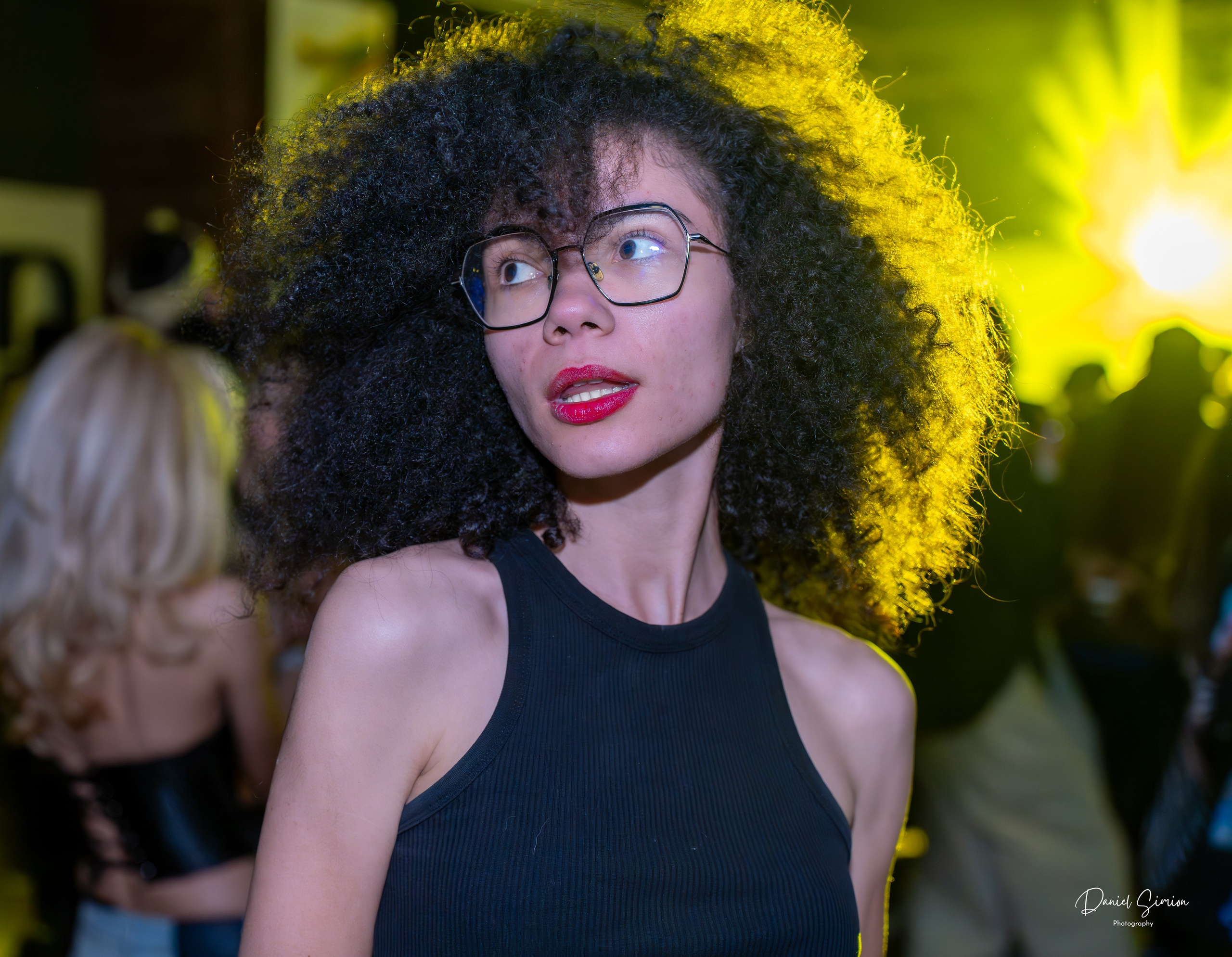 Young woman with curly hair and glasses at an indoor event with yellow backlighting.