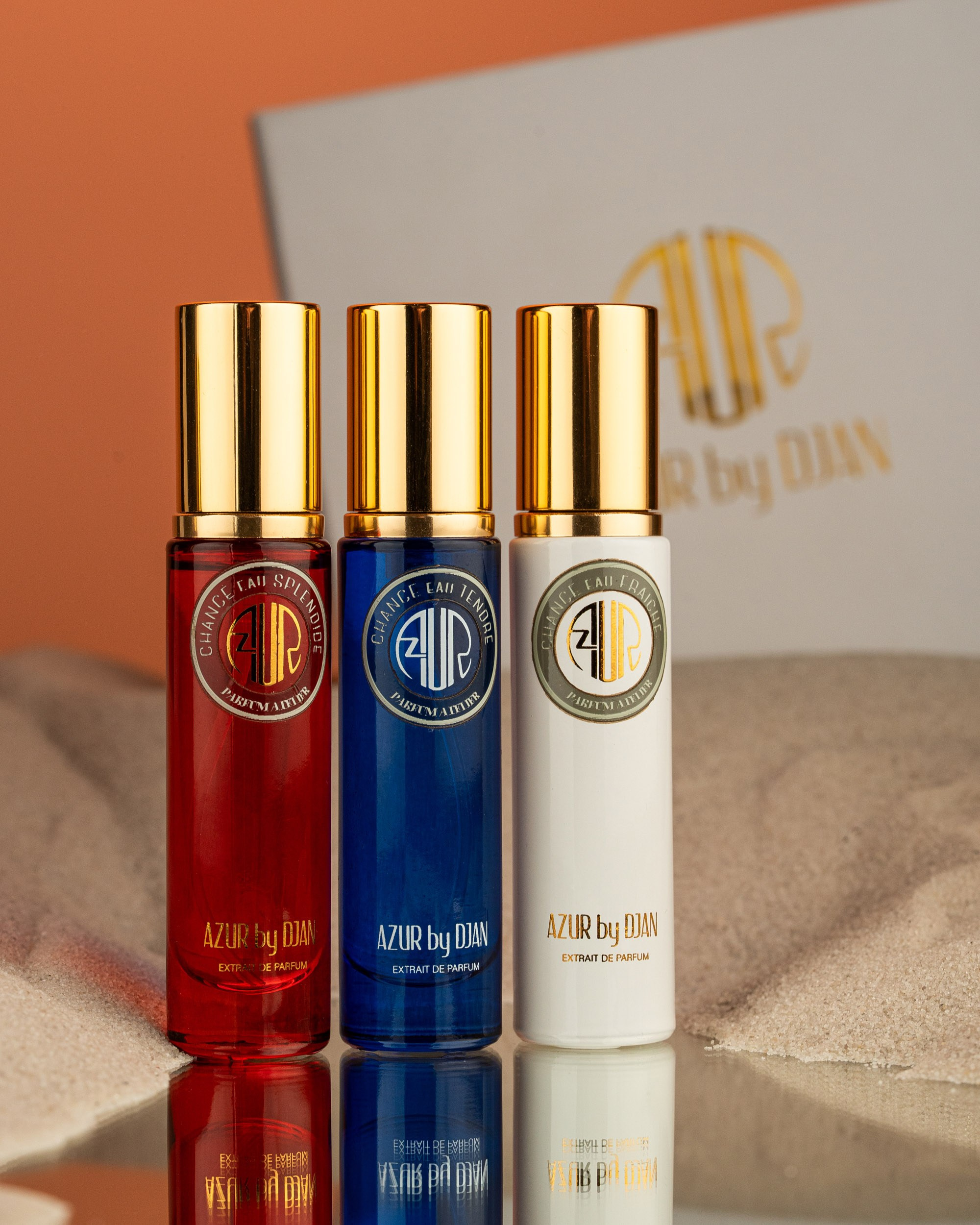 AZUR by DJAN brendinə aid üç kiçik, silindrik ətir şüşəsi açıq, qumlu fona qarşı əks etdirici səth üzərində düzülüb. Şüşələrin qızılı qapaqları və üzərində brend loqosu olan şəffaf etiketləri var. Onlar müvafiq olaraq qırmızı, mavi və ağ/şəffaf maye ilə doldurulub. Brendin ağ qutusu bulanıq arxa planda qismən görünür.