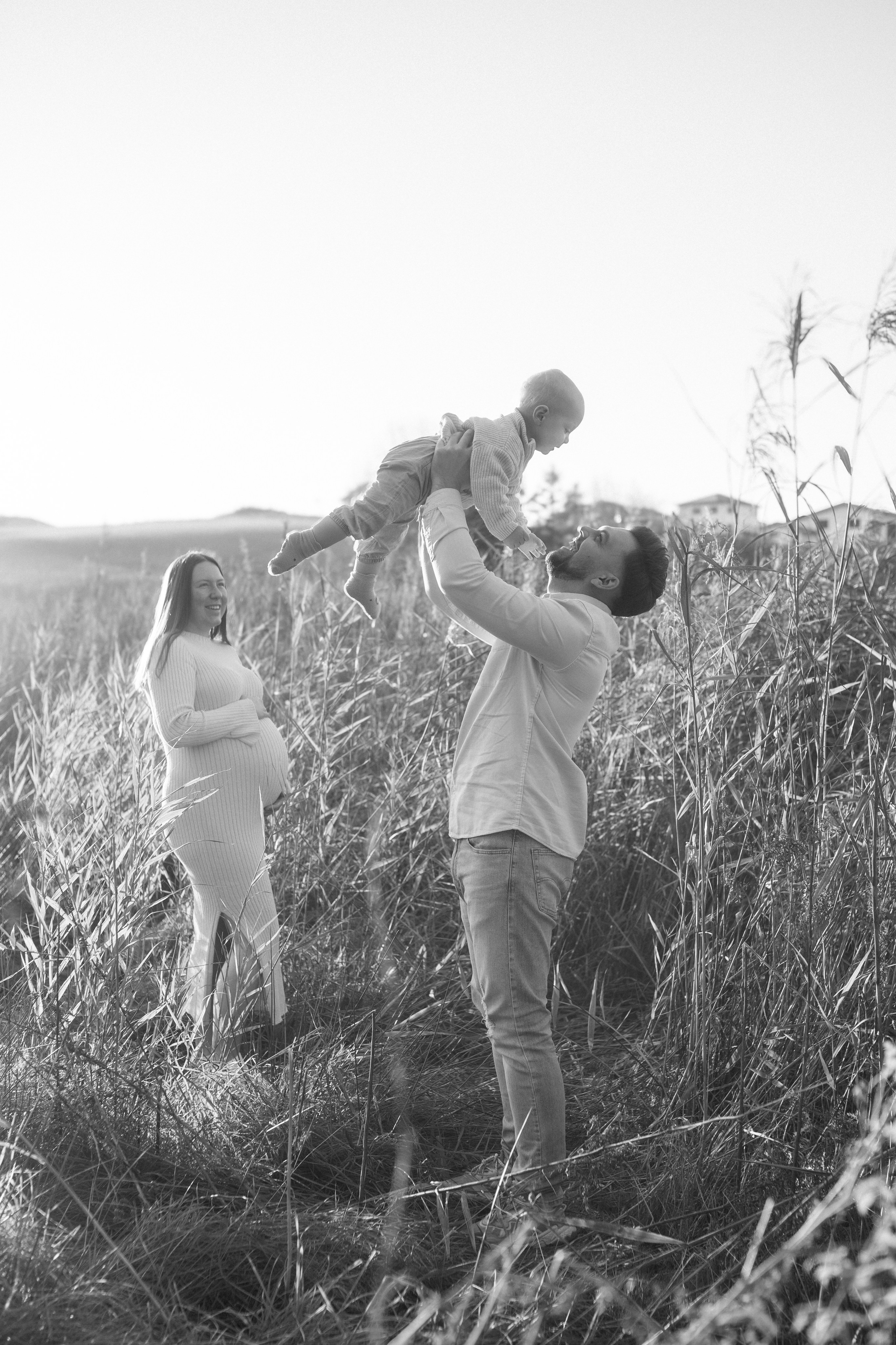 Glückliche Familie im Freien – die schwangere Mutter schaut liebevoll, wie ihr Mann den Sohn in die Luft hebt