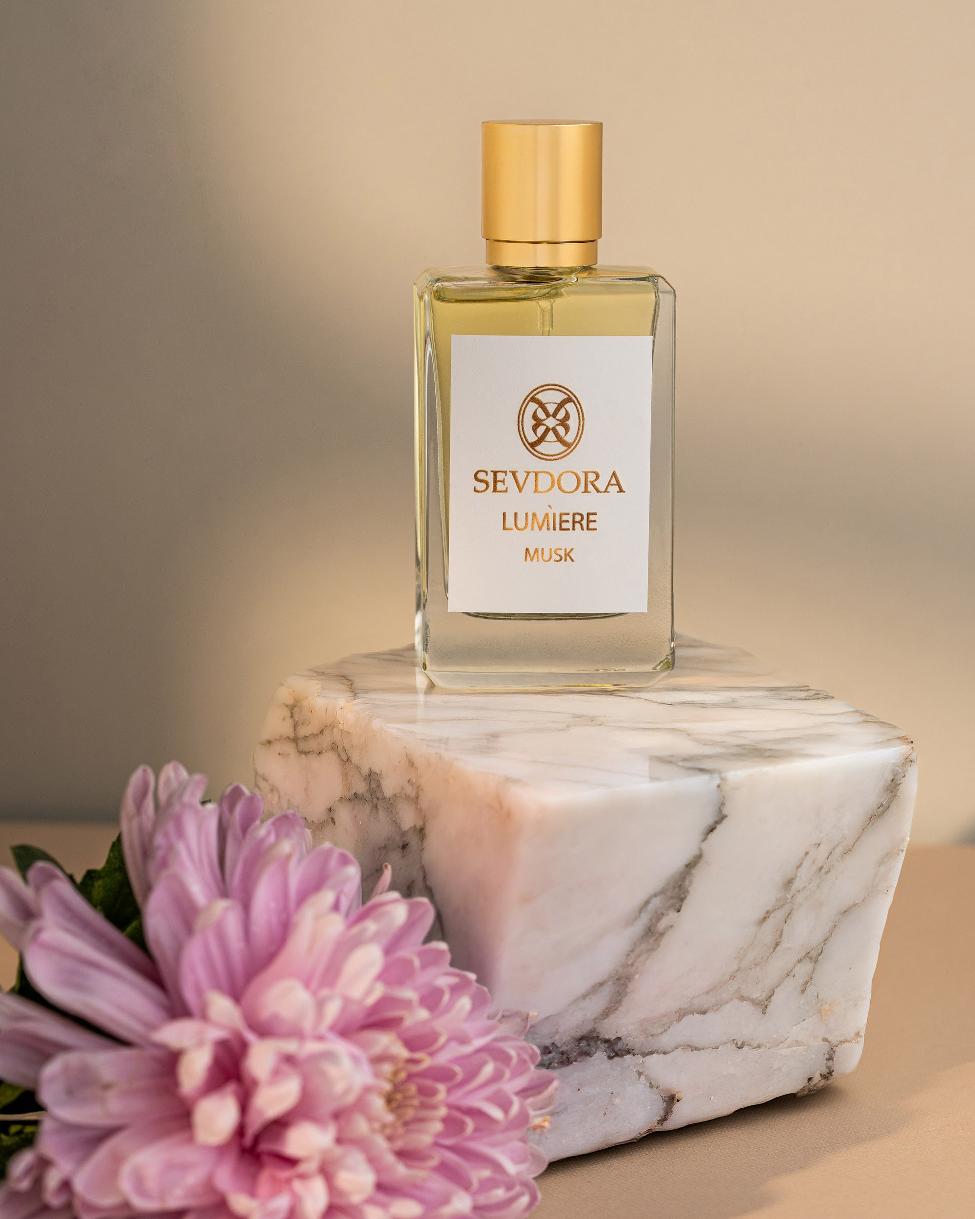 "SEVDORA LUMIERE MUSK" yazısı olan şüşə ətir butulkası ağ mərmər bloku üzərində dayanır. Ön planda iri, çəhrayı rəngli xrizantema çiçəyi bulanıq şəkildə görünür.