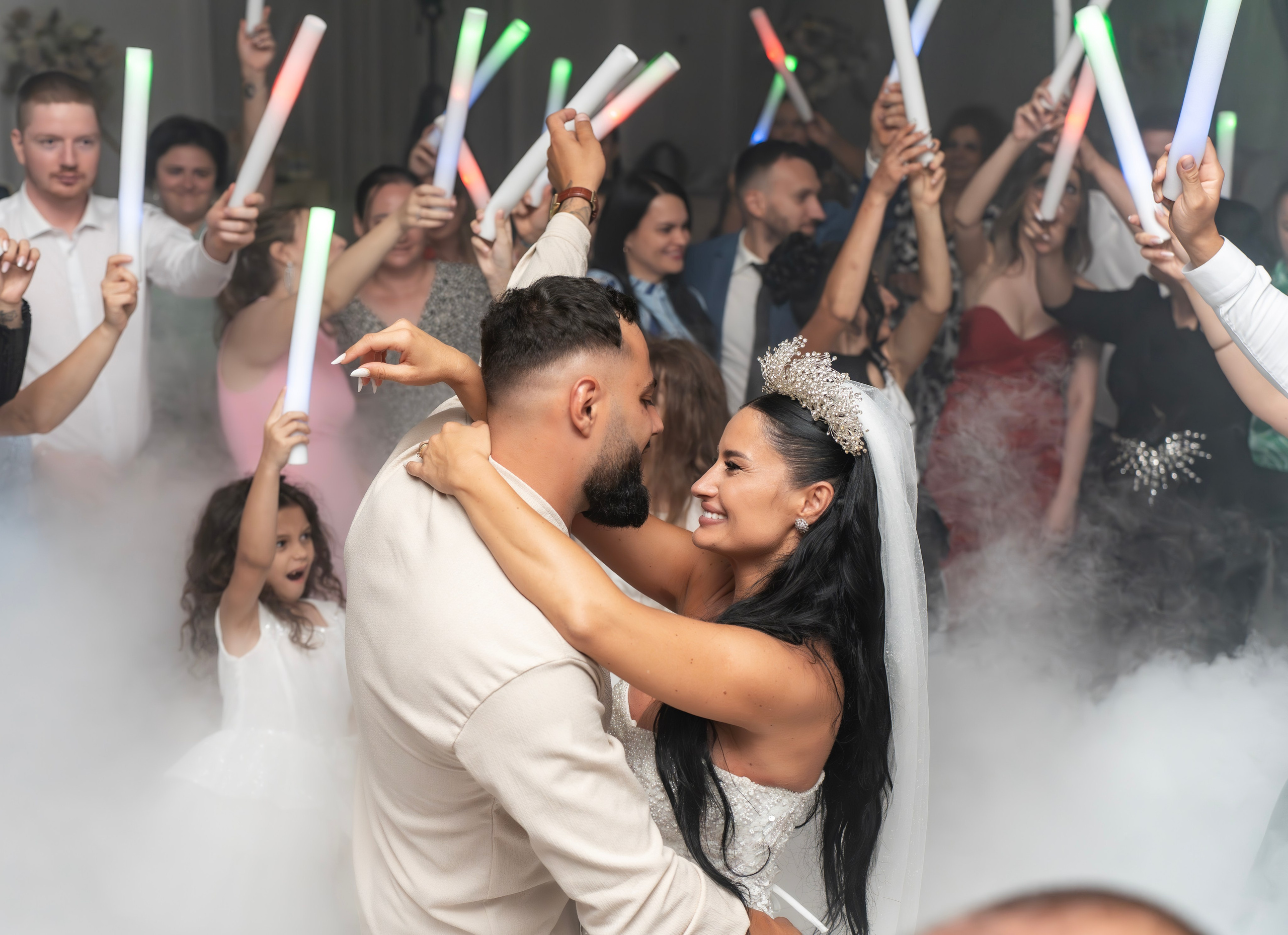 Bride, groom, and two guests cutting a white wedding cake on a glass table at a wedding reception.