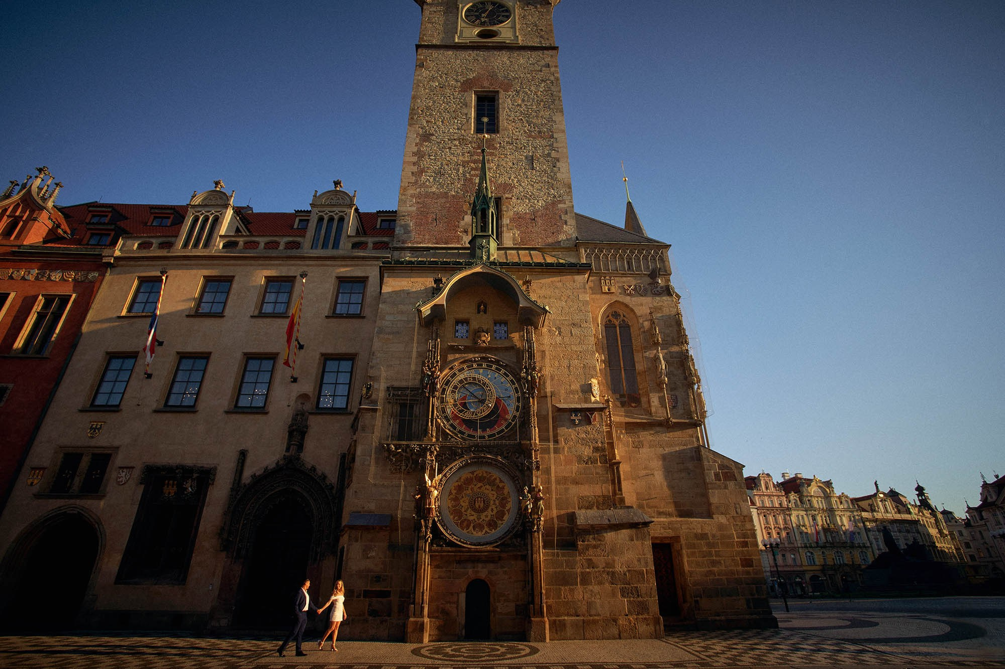 A young California couple standing under the Astronomical Clock as sunrise fills Old Town Square.