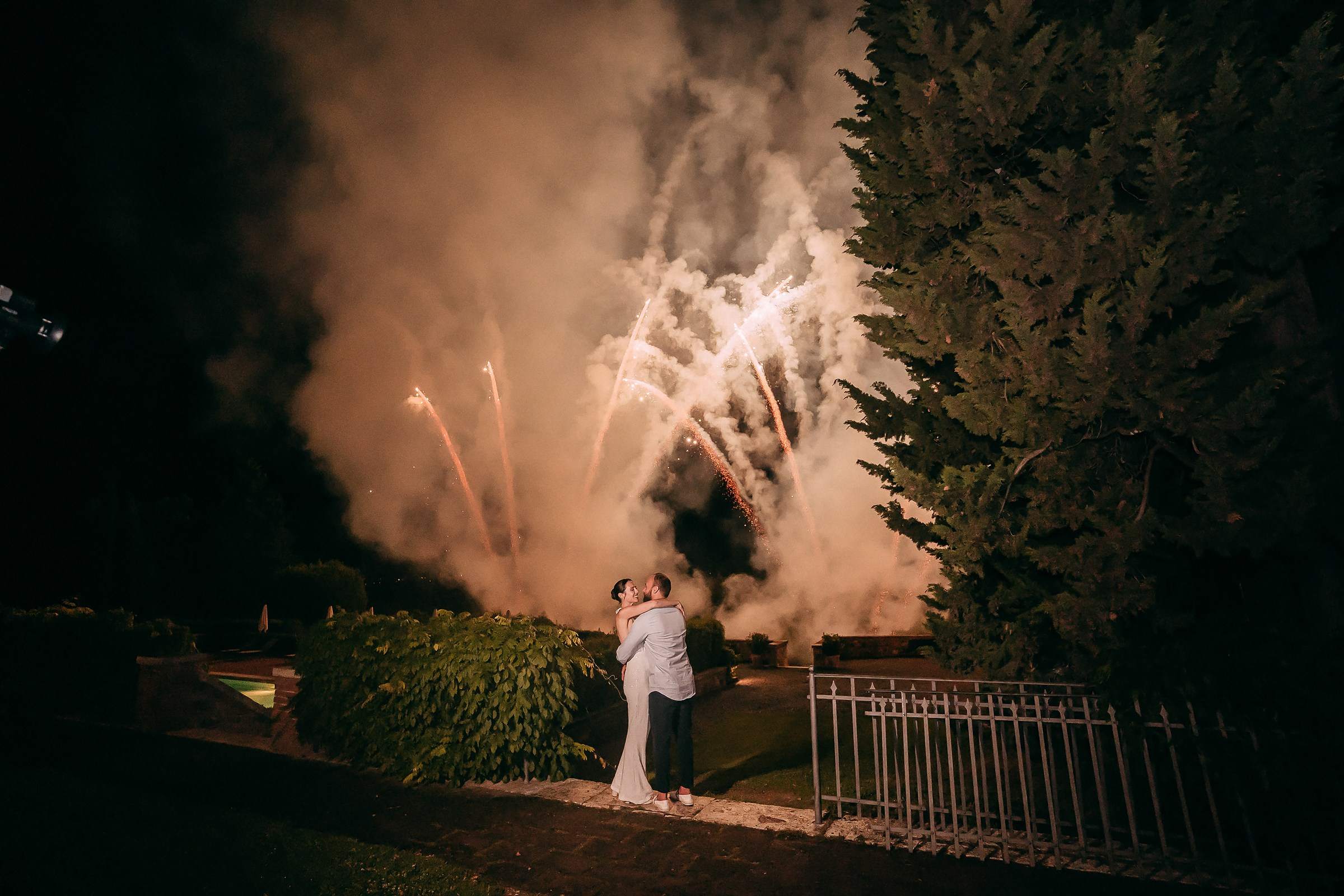 A couple shares a romantic kiss under a dramatic night sky illuminated by vibrant fireworks, with a faint haze from the smoke adding to the atmosphere.