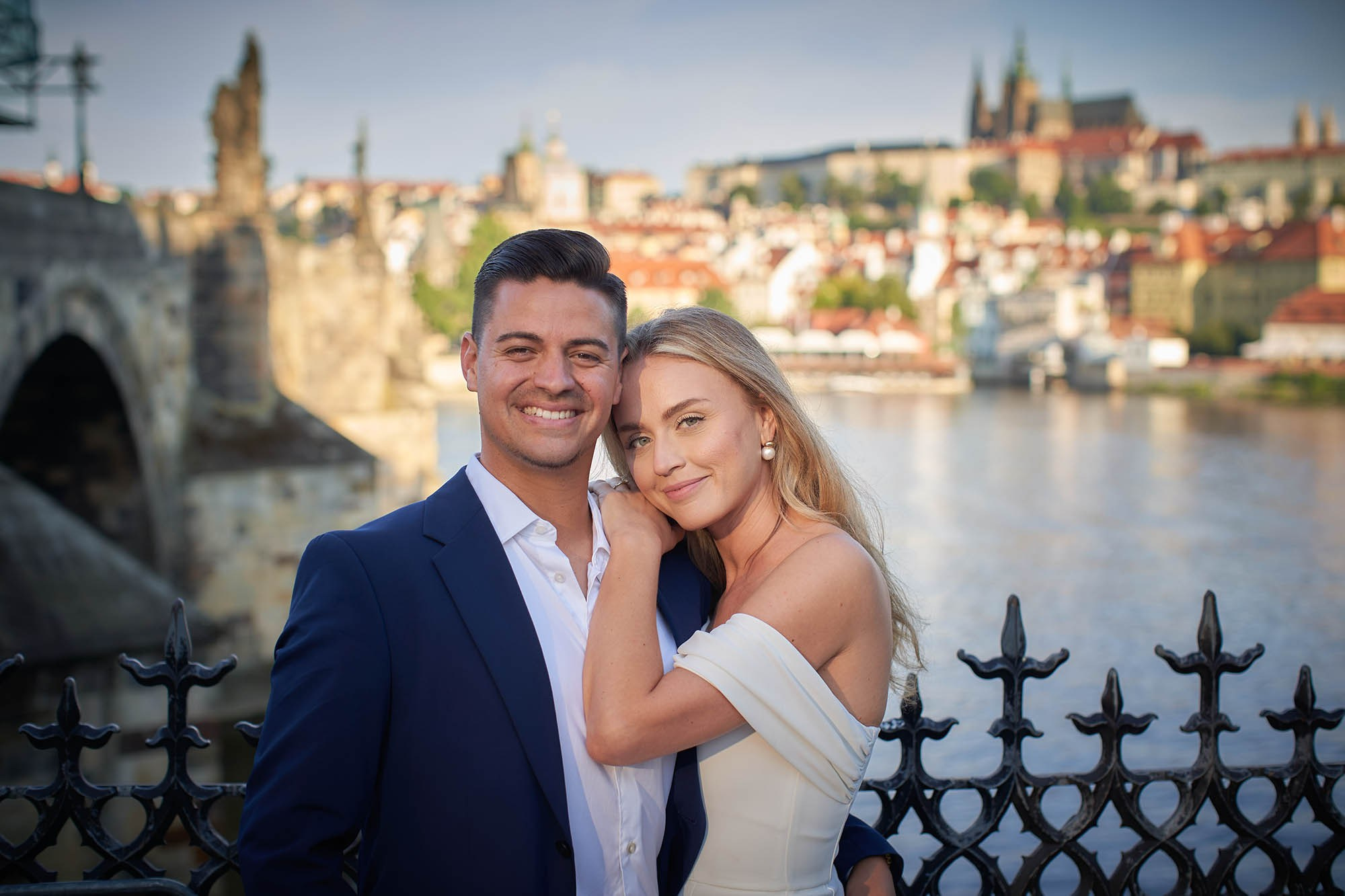 Smiling engaged couple portrait with dramatic Prague Castle sunrise skyline background.