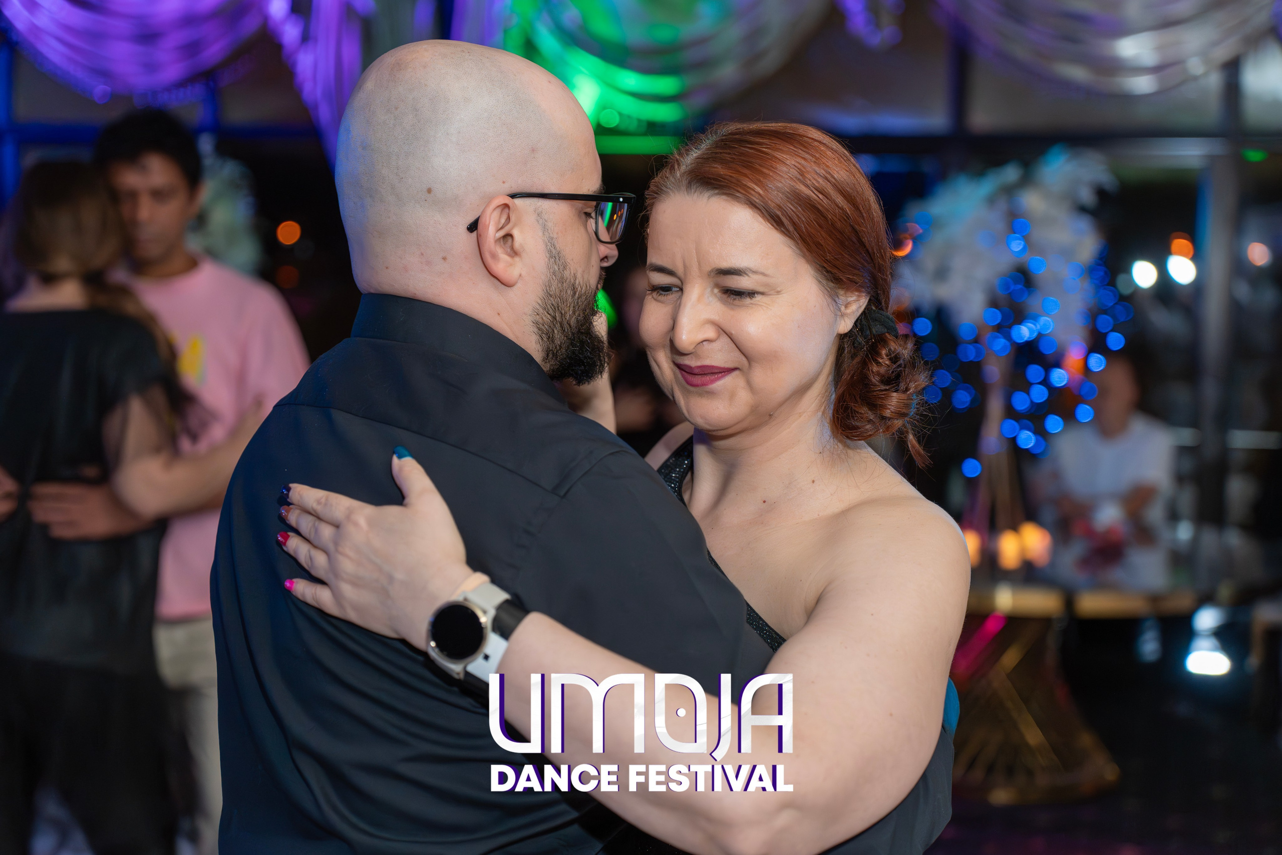 Man and woman dancing closely in a softly lit room with blurred background figures.