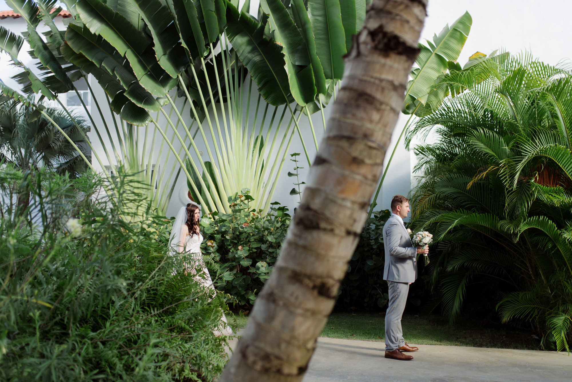 The groom in a light suit with a bouquet of flowers stands in the shade of palm trees.
