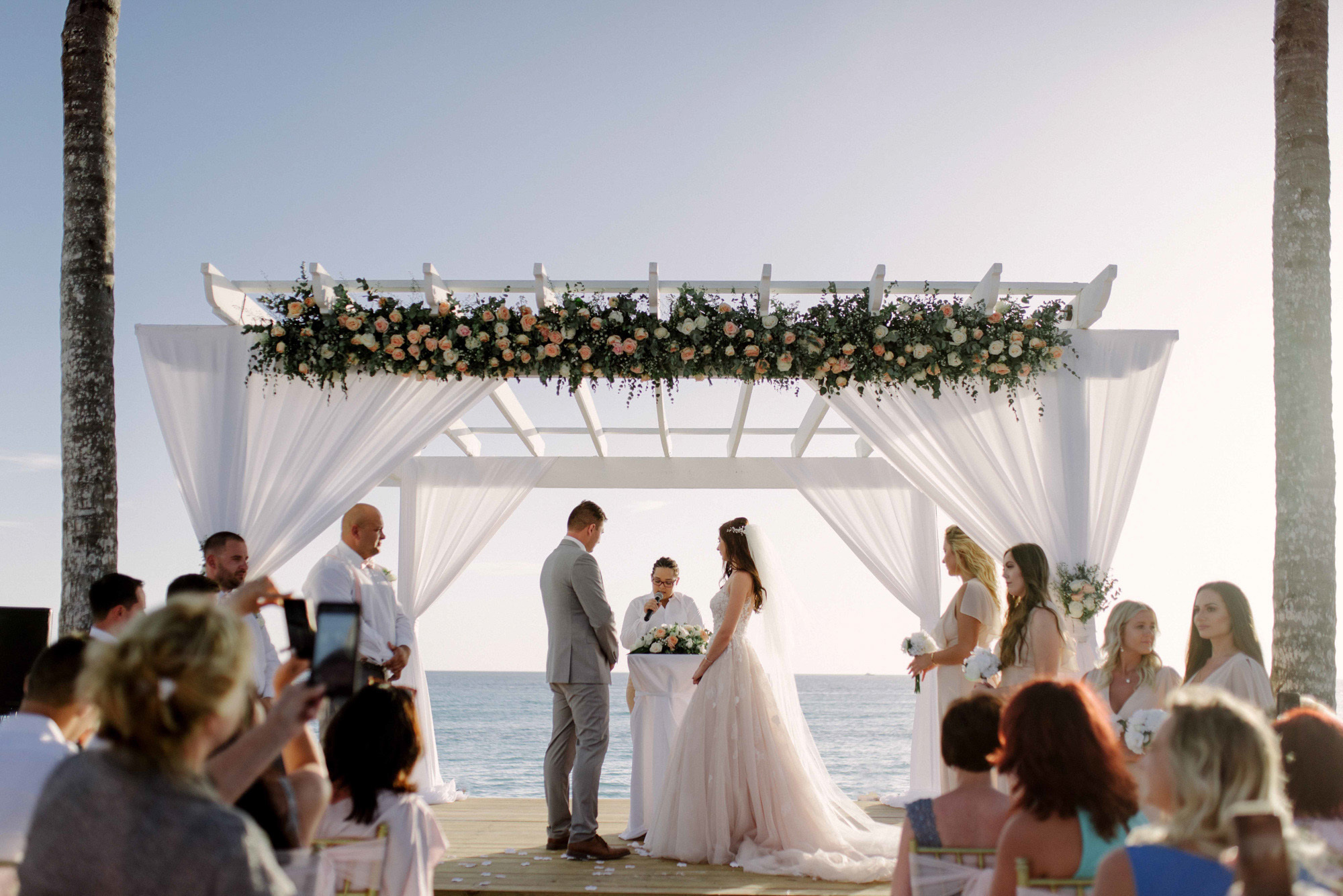 The bride and groom stand at the altar in front of the priest surrounded by guests on the background of the sea.