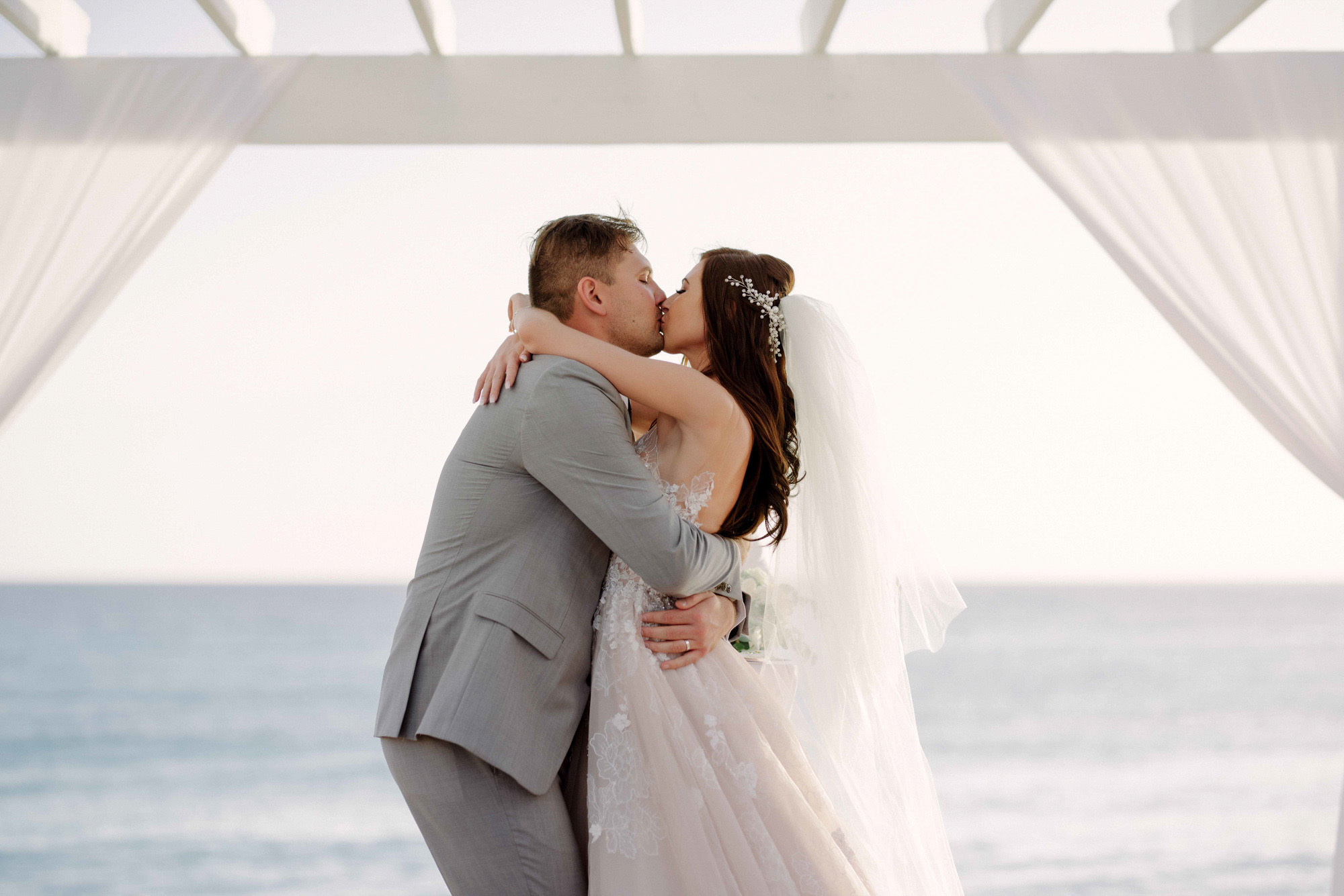 Bride and groom under the altar with white curtains hug and kiss each other on the background of the sea.