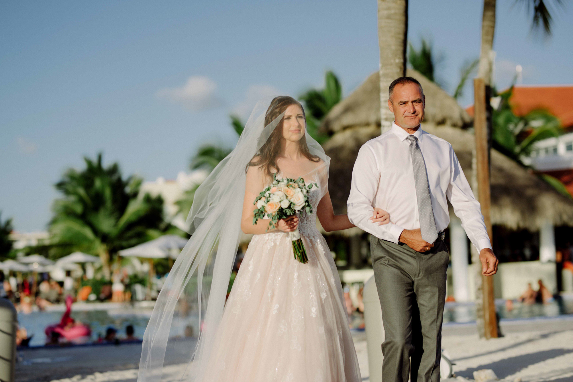 Bride and dad walk down the aisle in front of wedding tables, palm trees and beach houses.