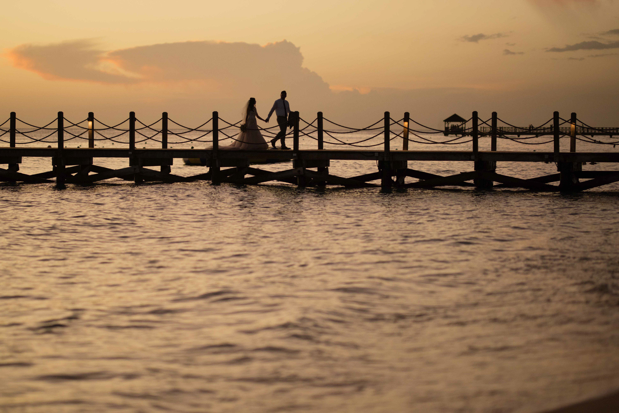 Bride and groom walk on wooden pier with sea background.