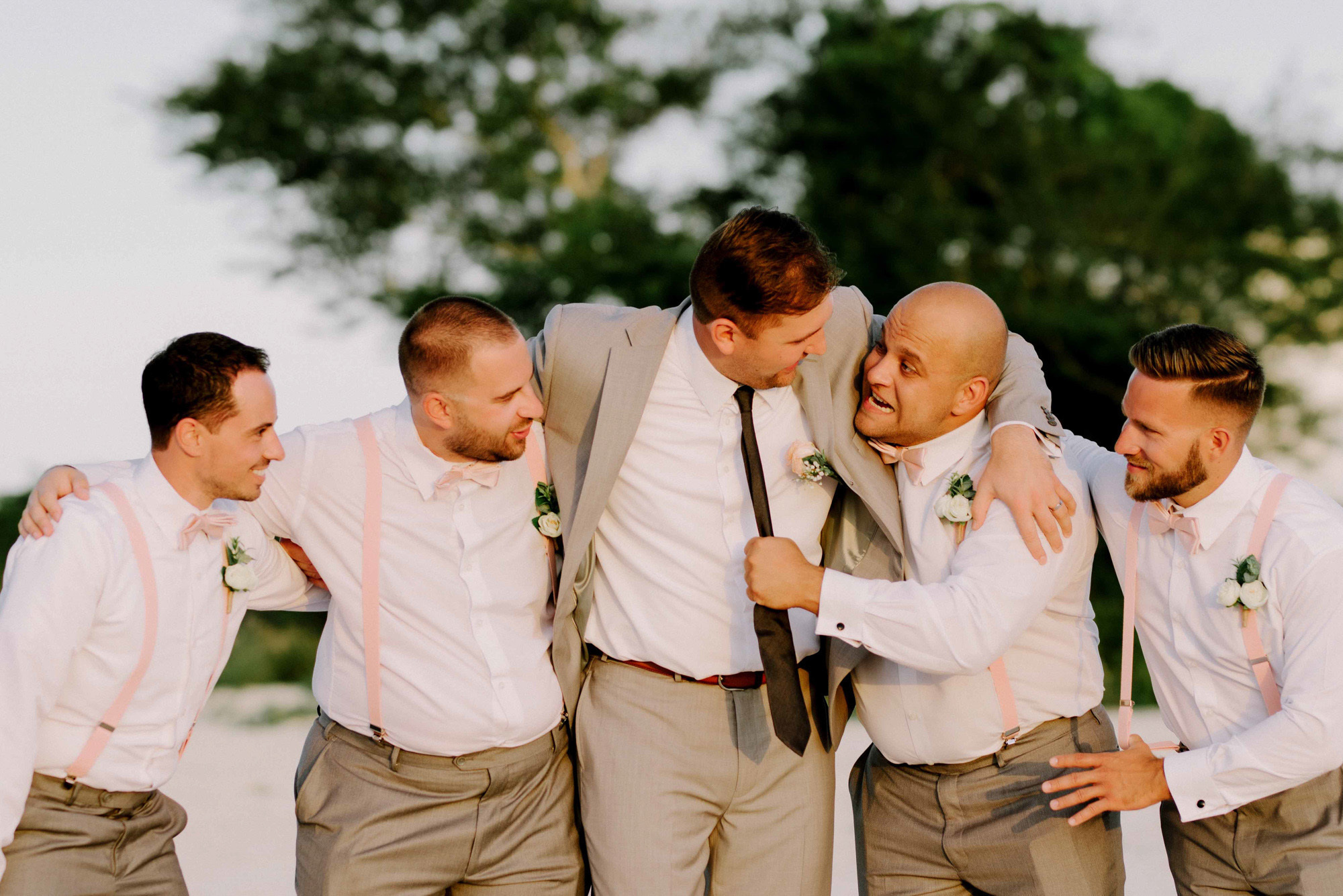 The groom stands on the beach, hugging friends against the background of green trees.