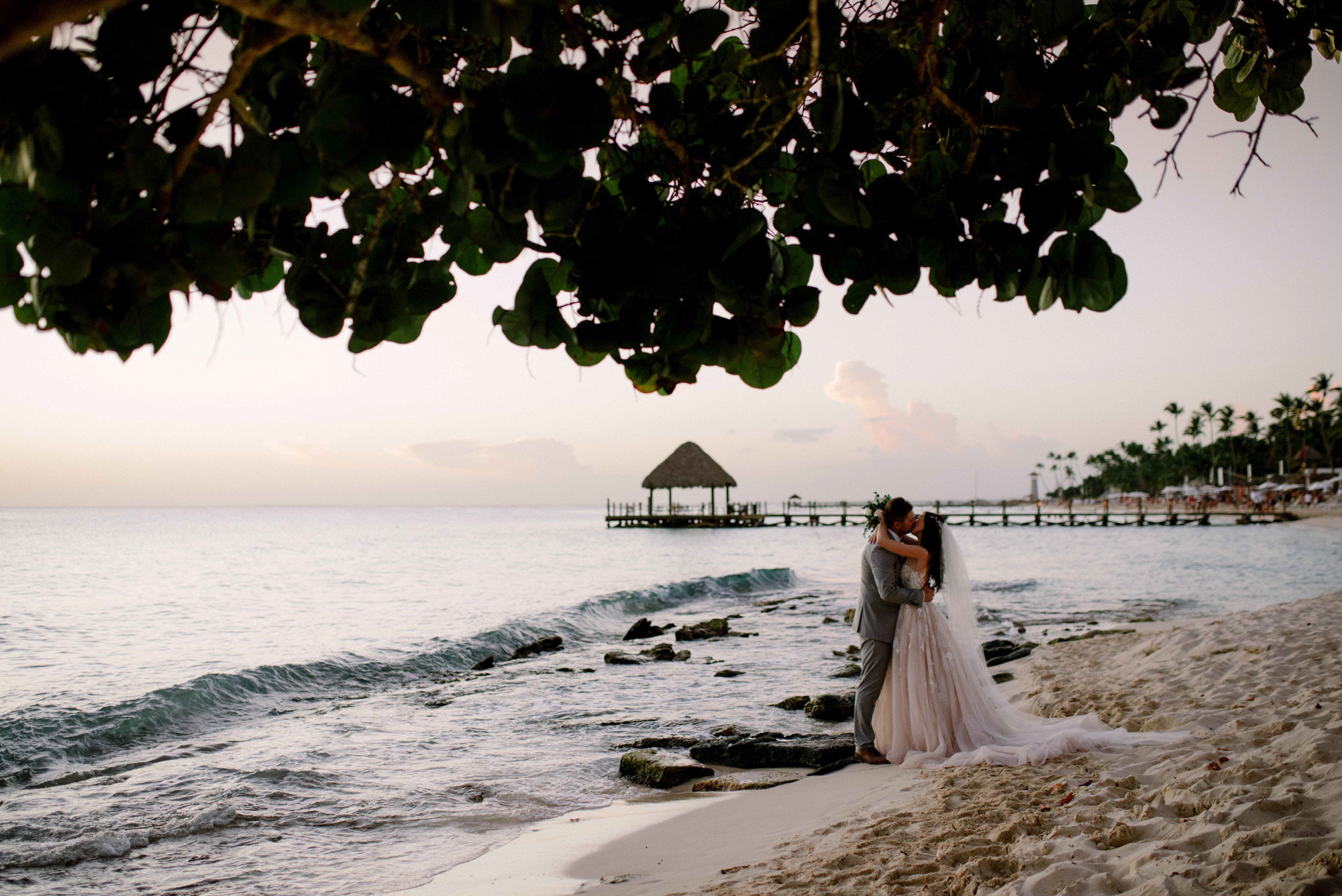 Bride and groom kiss at the edge of the ocean on the background of a beach with palm trees and a beach house.