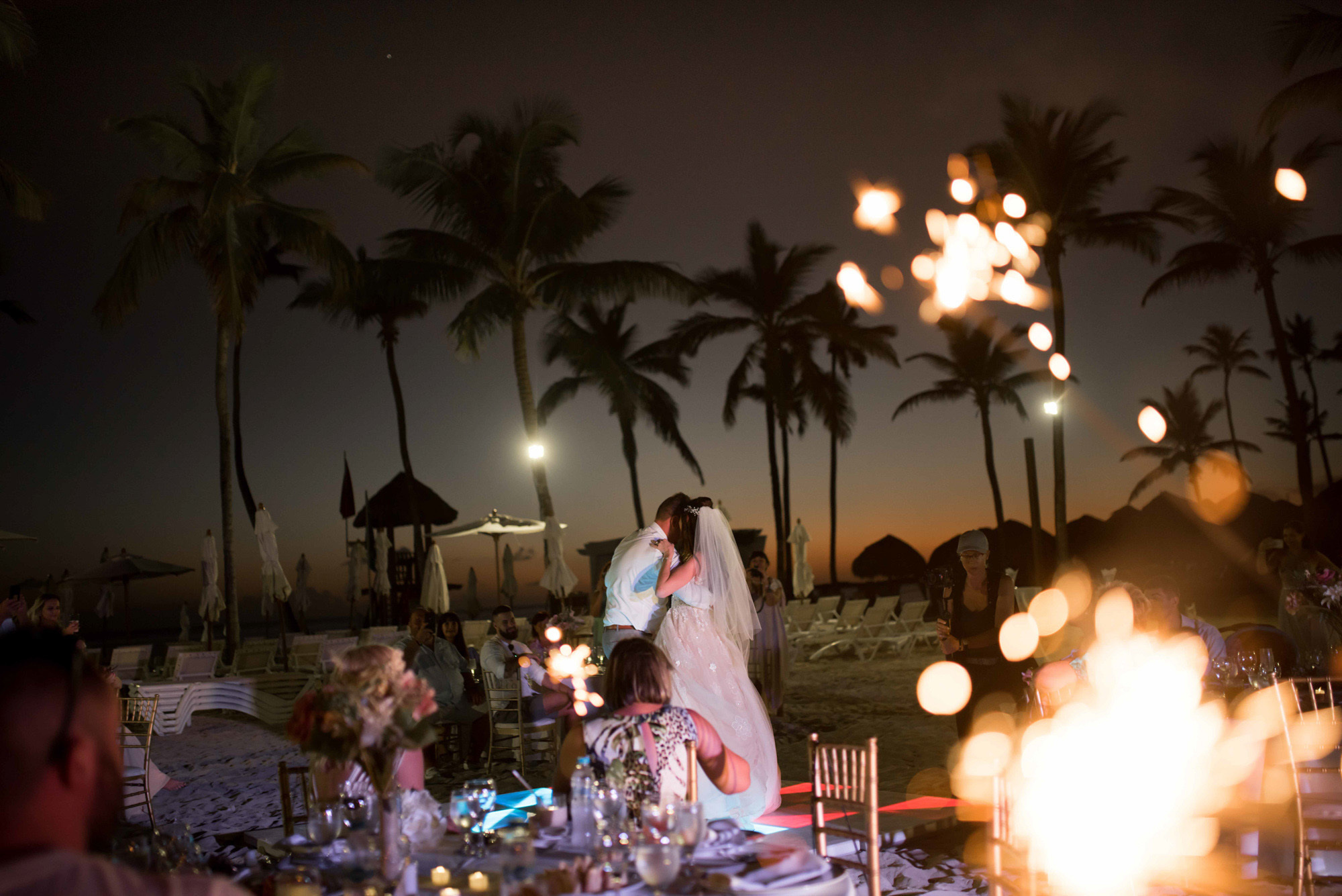 From behind the wedding tables, the bride and groom can be seen dancing against the background of the night sky and palm trees.
