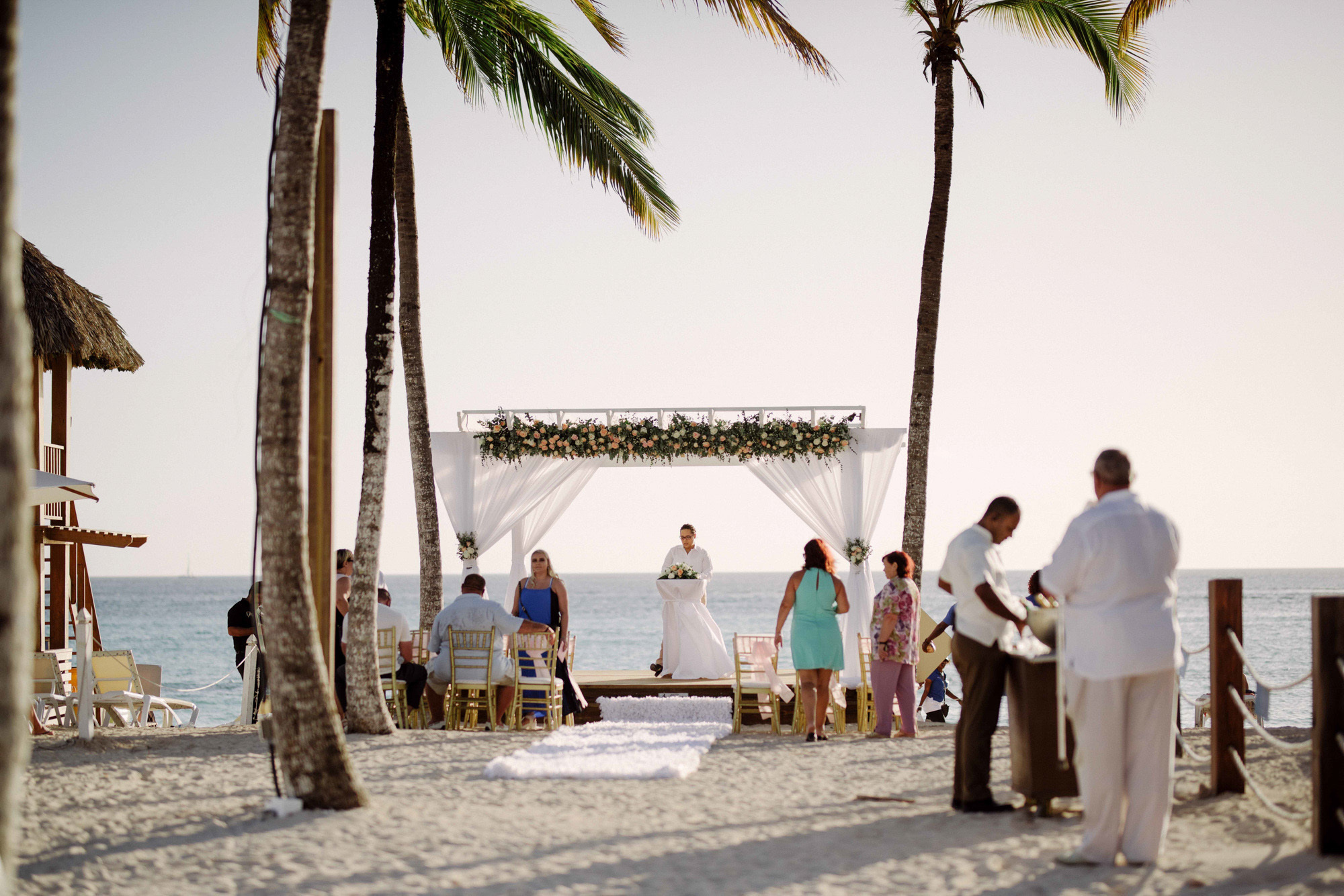 At the altar with white curtains on the background of the sea beach stands a priest and several guests.