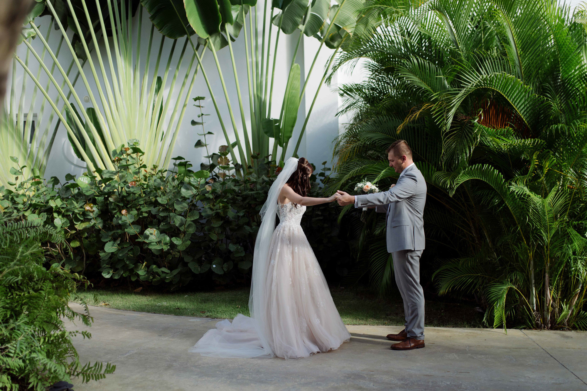 Among the green oasis, the groom holds the bride's hand, admiring her outfit.
