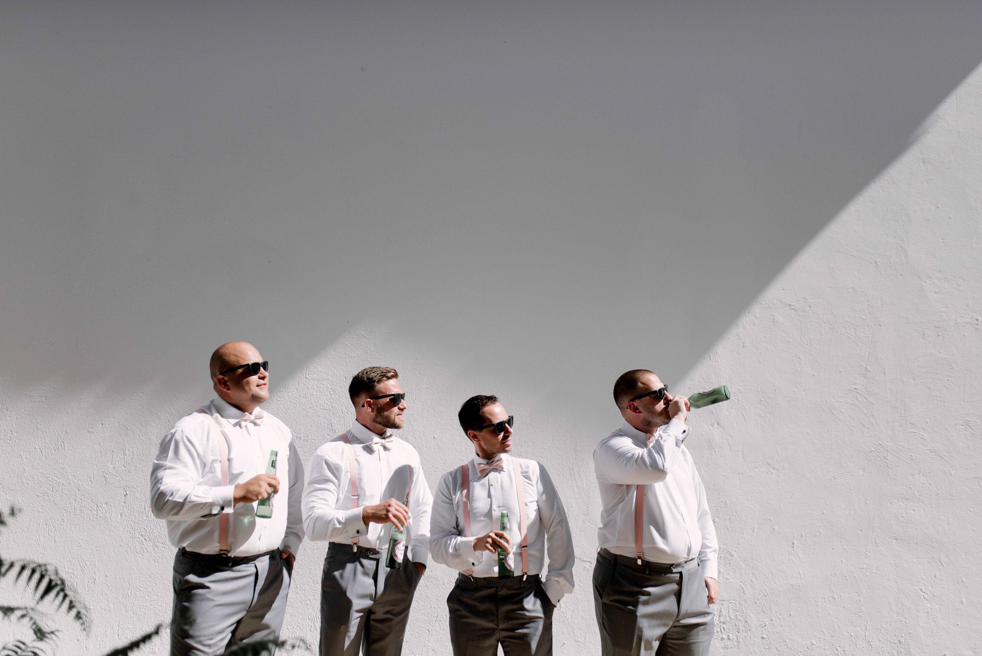 The groom drinks champagne from a bottle, and three of his friends hold champagne glasses in their hands. All of them are wearing light trousers with suspenders, white shirts and dark sunglasses against a white background of buildings.