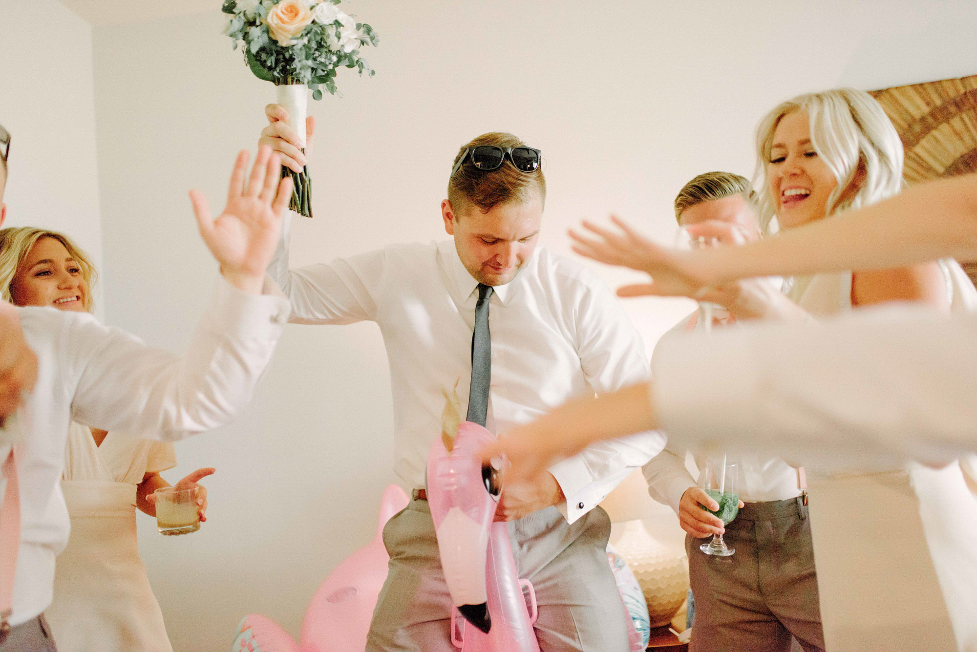 Groom with a bouquet of flowers jumps on an inflatable flamingo.