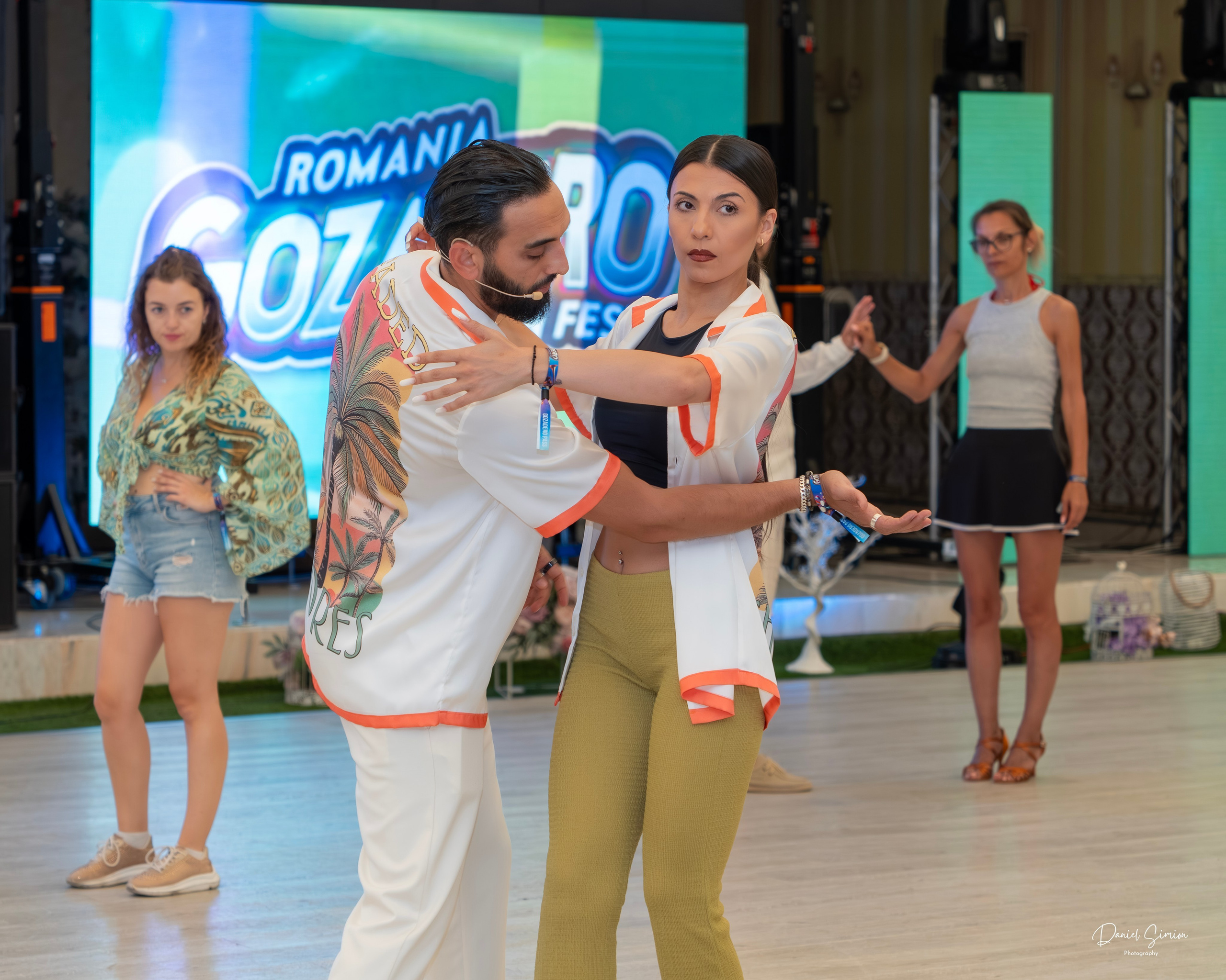 Dance instructor guiding a female student in close partner dance with people observing in background.