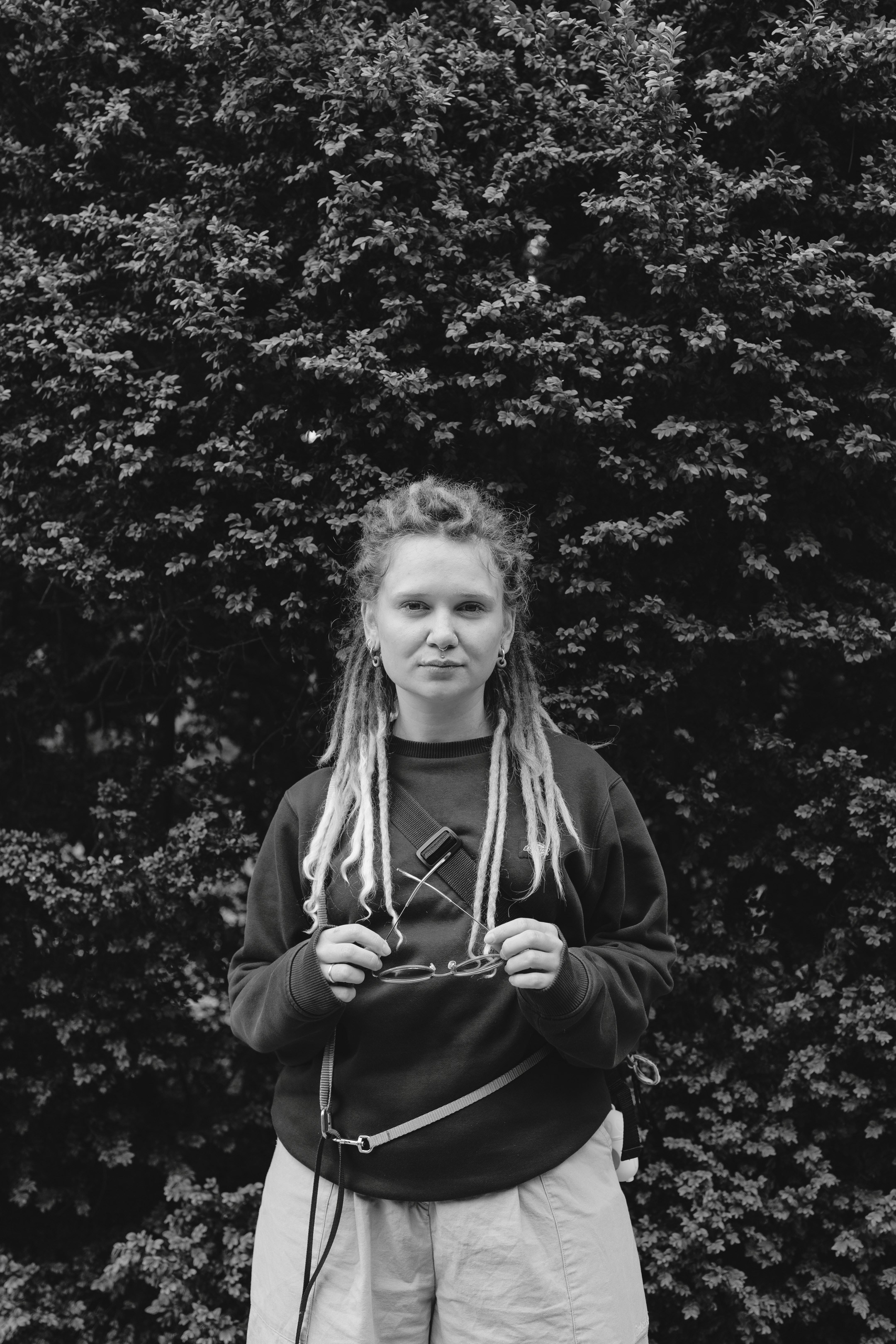 Чёрно-белый портрет Юли с дредами на фоне зелени, задумчивый взгляд.