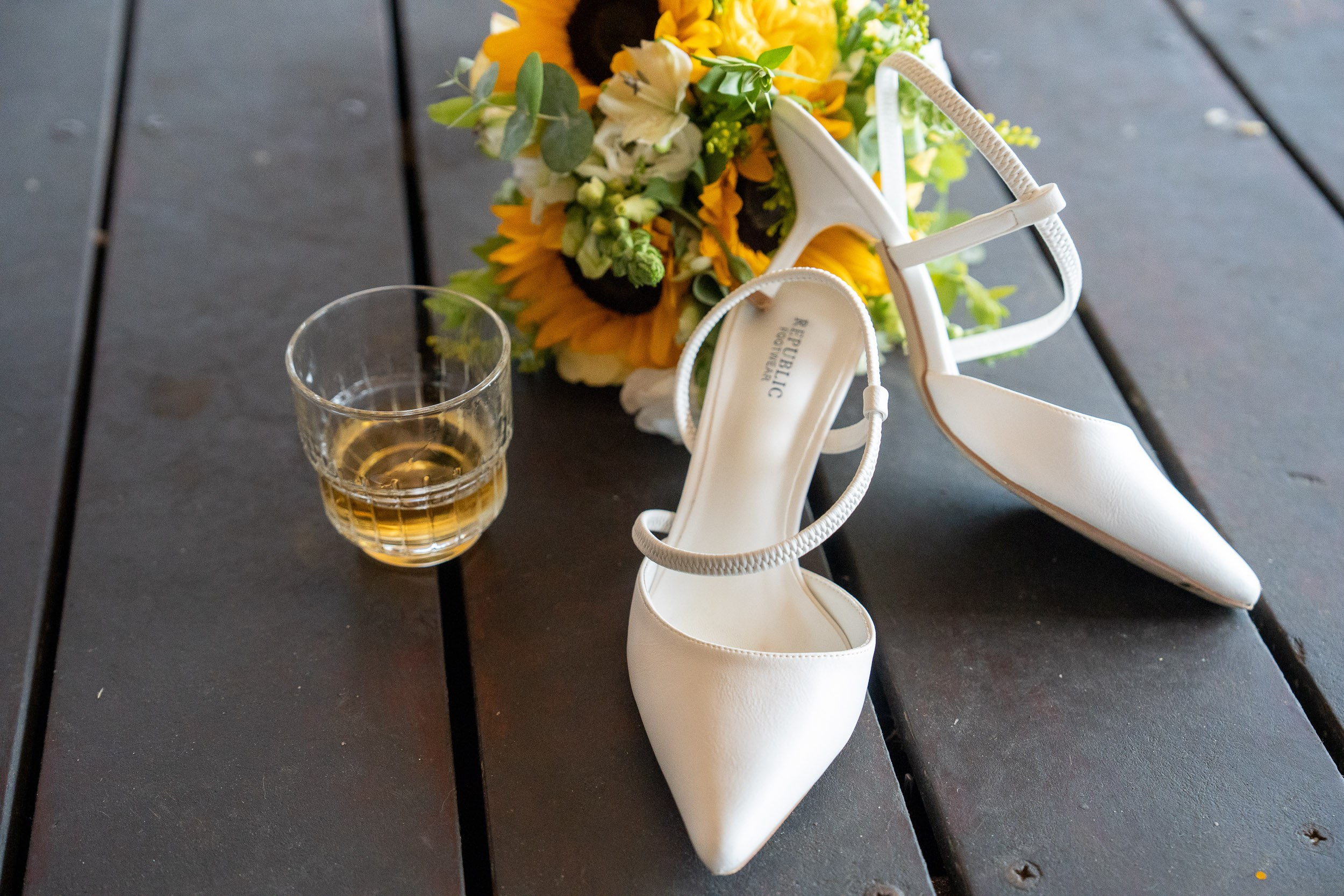 Wedding shoes, a sunflower bouquet, and a glass of whisky arranged together