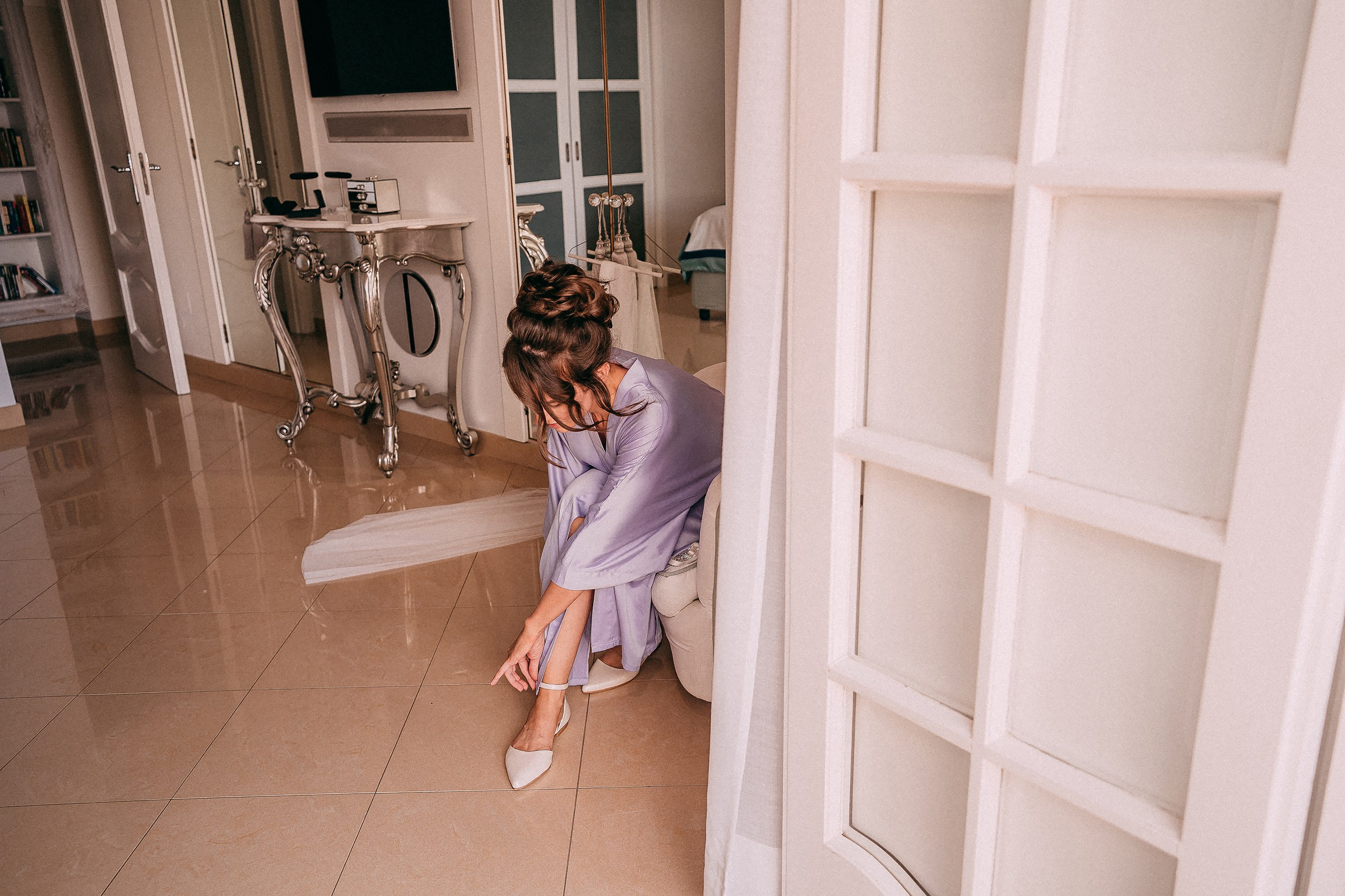 A bride in a lavender robe kneels to adjust her heels inside Villa Bohème, surrounded by bright, luxurious decor.
