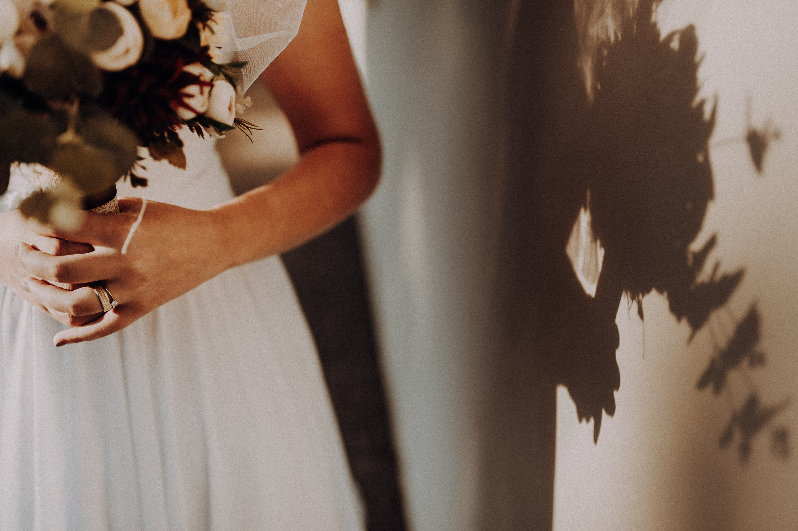 Umbra buchetului de mireasa pe perete avand in prim plan verighetele de pe mana miresei.