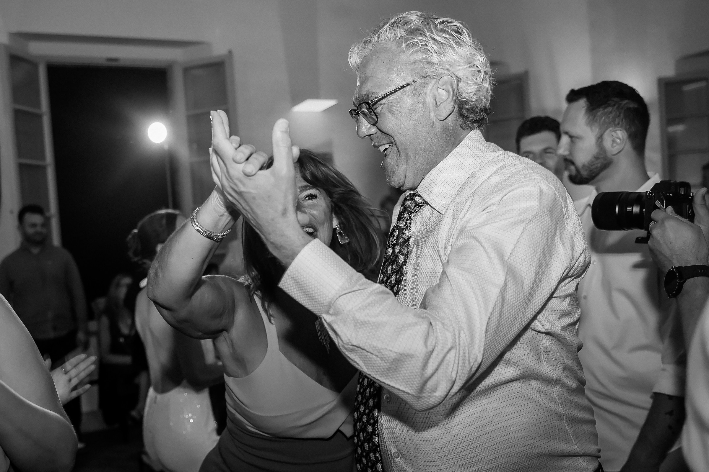 A black-and-white photo capturing a joyful moment on the dance floor as two guests energetically dance together, smiling and holding hands.