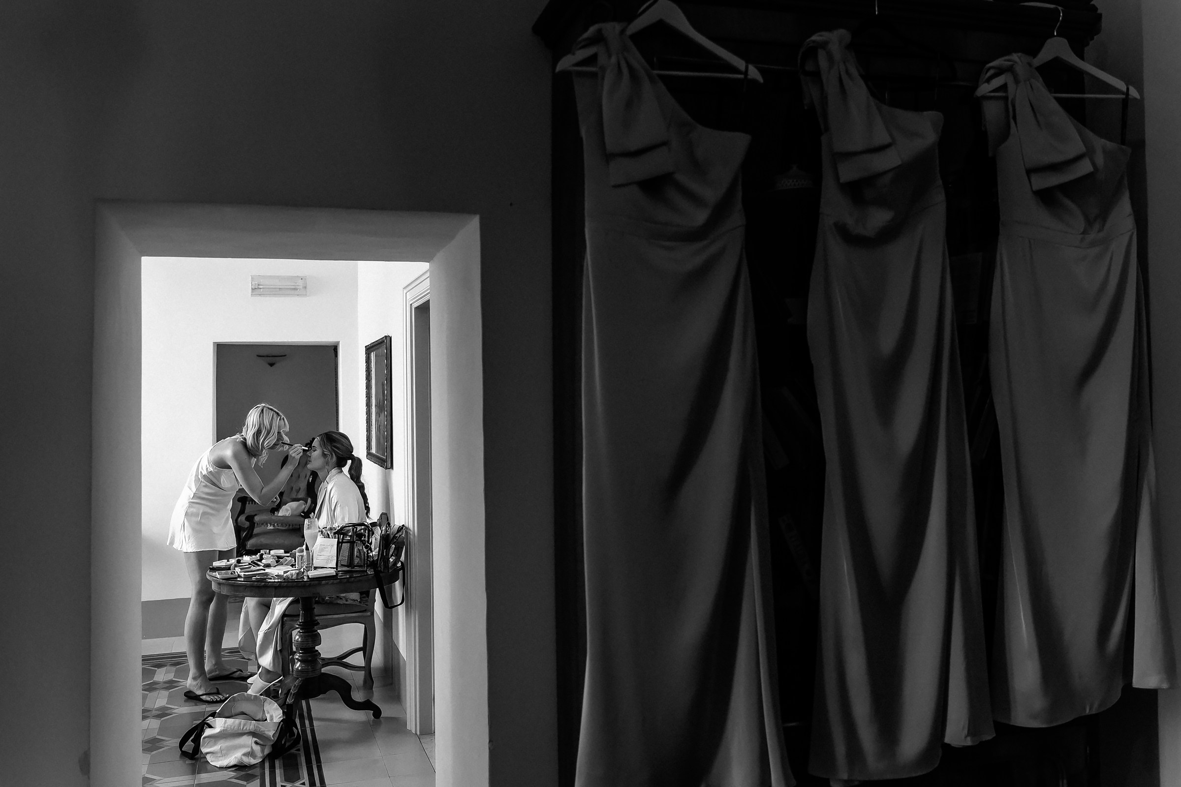 A black-and-white capture of a bride being prepared for her special day, as her dress hangs elegantly in the doorway.