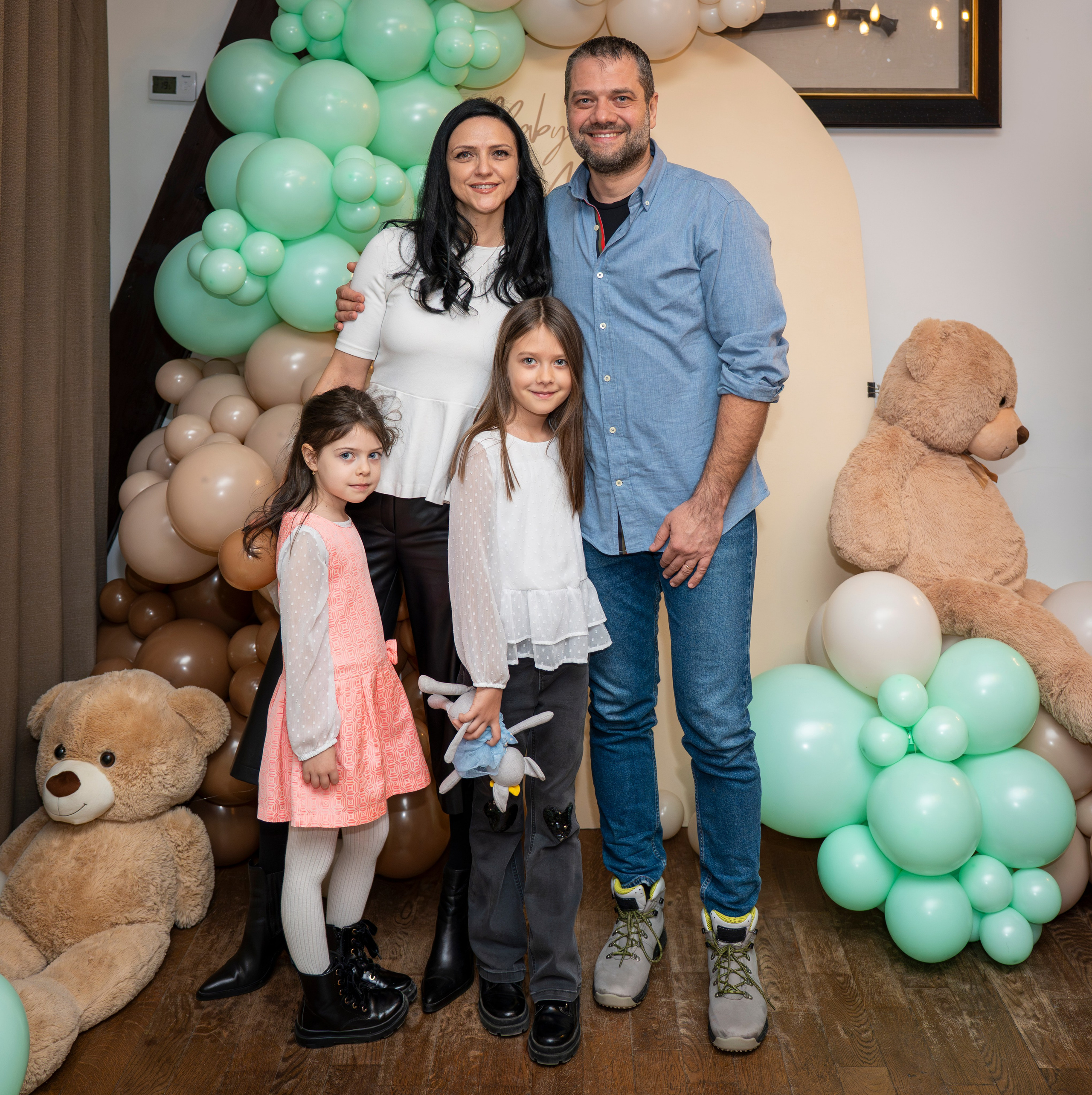 Family of four posing indoors with beige and mint green balloons and two large teddy bears.