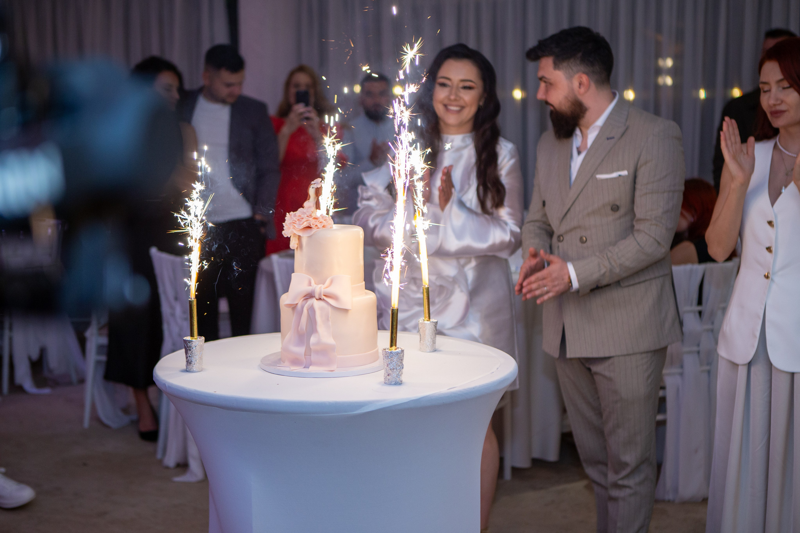 A couple clapping near a two-tiered white cake with sparklers on a decorated table during a celebration.