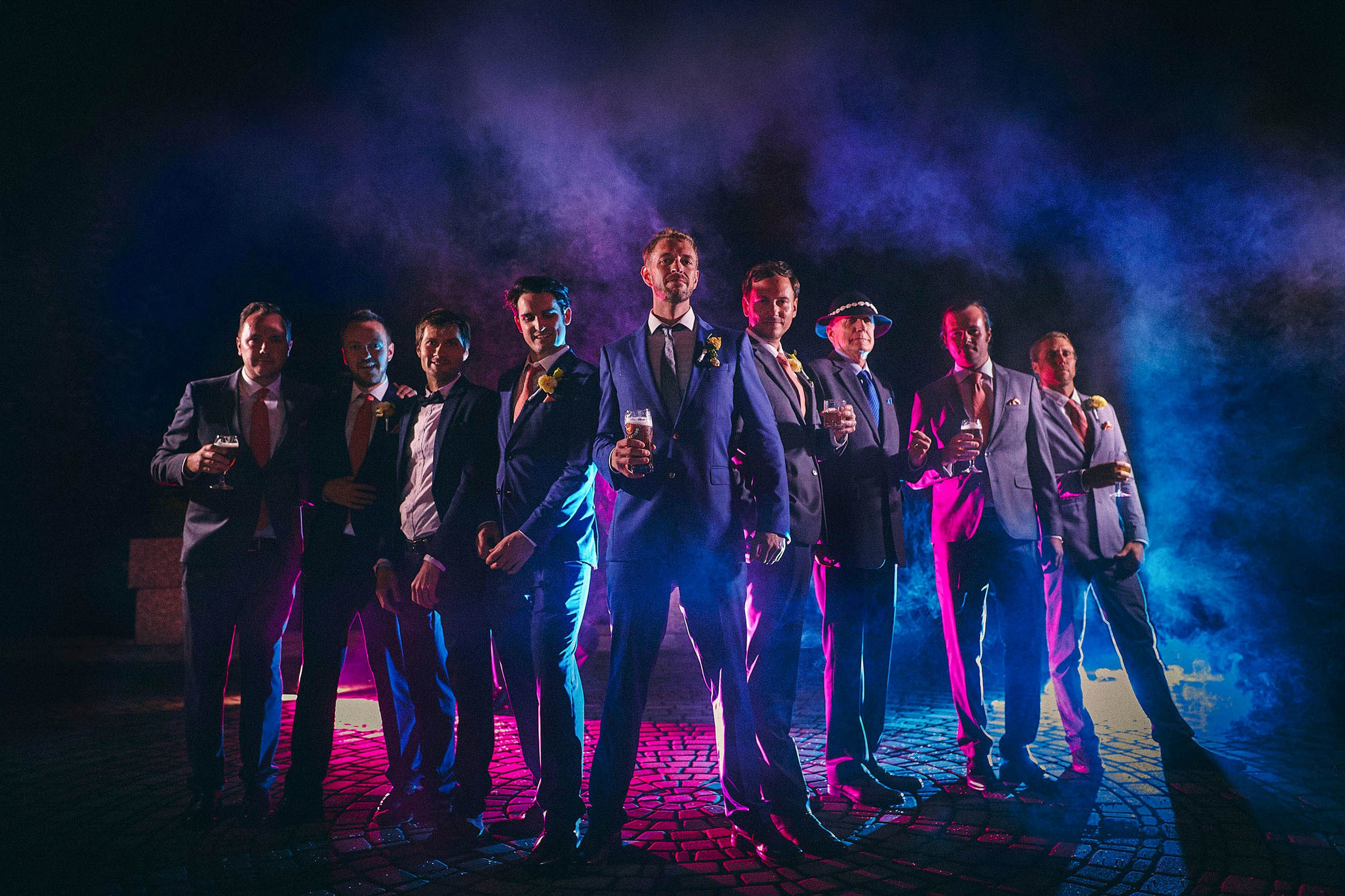 The best men, many holding beers, pose for a nighttime portrait with blue and purple smoke grenades.
